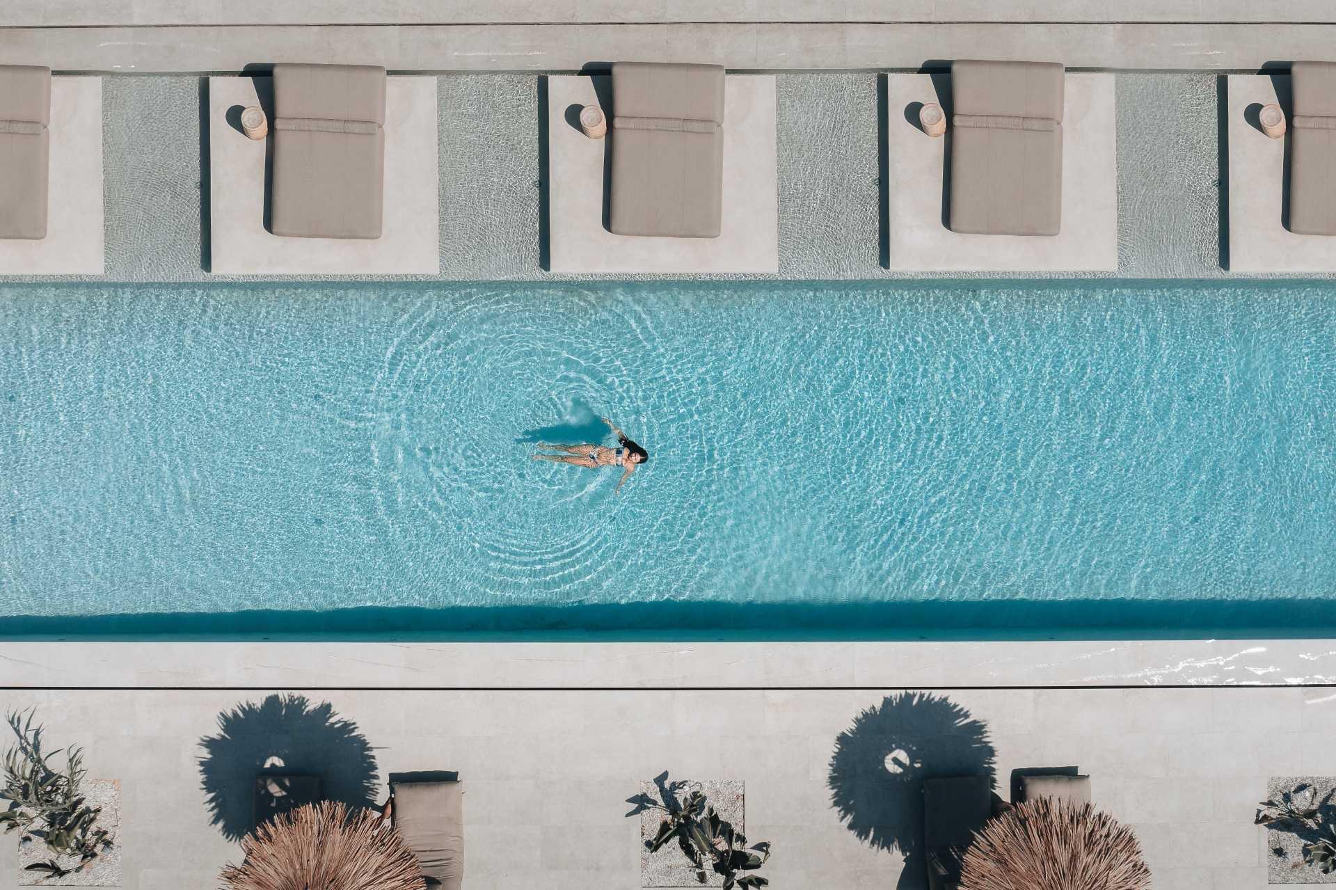 A pool seen straight from above, with sun loungers along both sides and a swimmer leaving ripples near the centre.