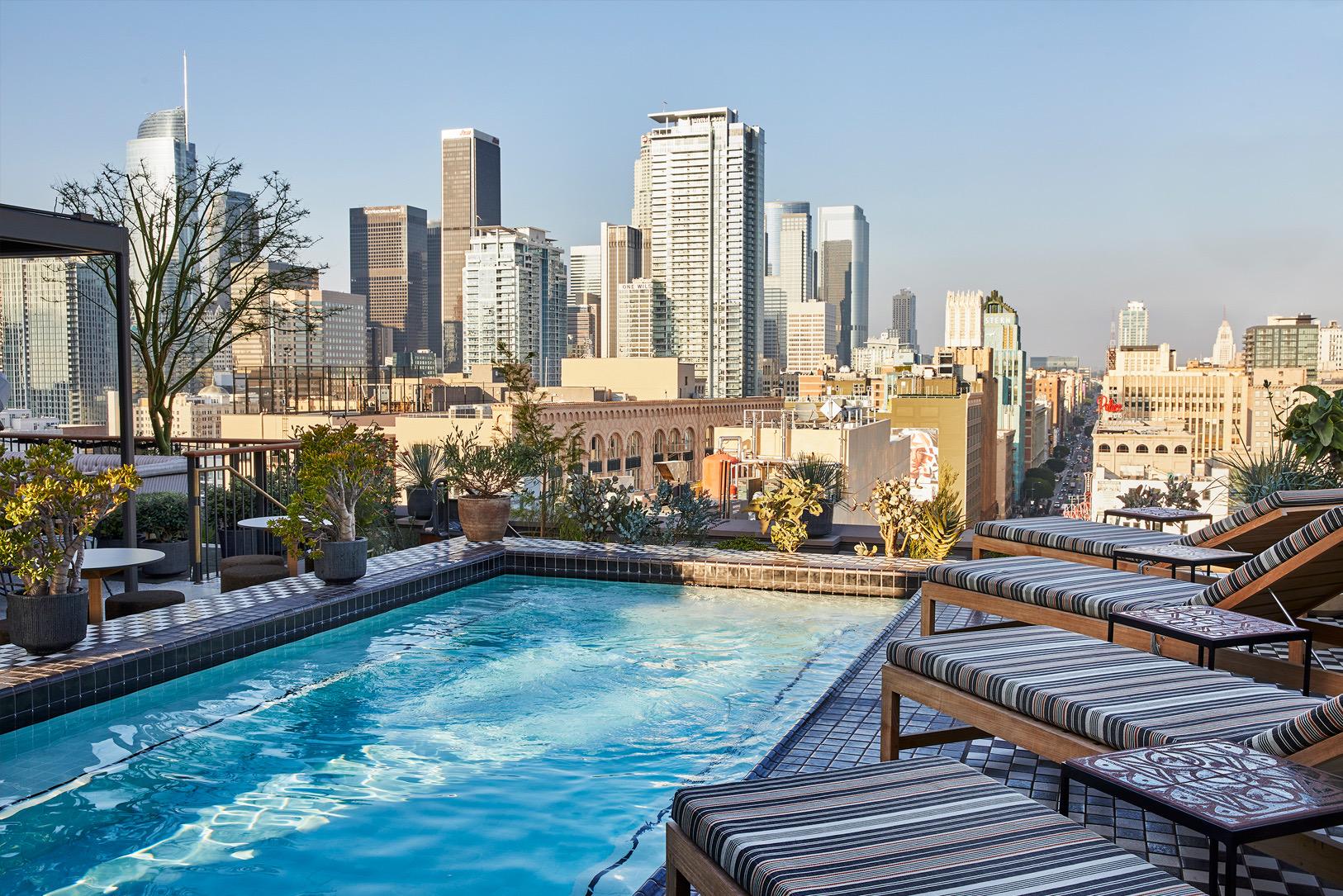 A rooftop pool lined with striped loungers, planters of palms and agave, and the downtown Los Angeles skyline beyond the railing.