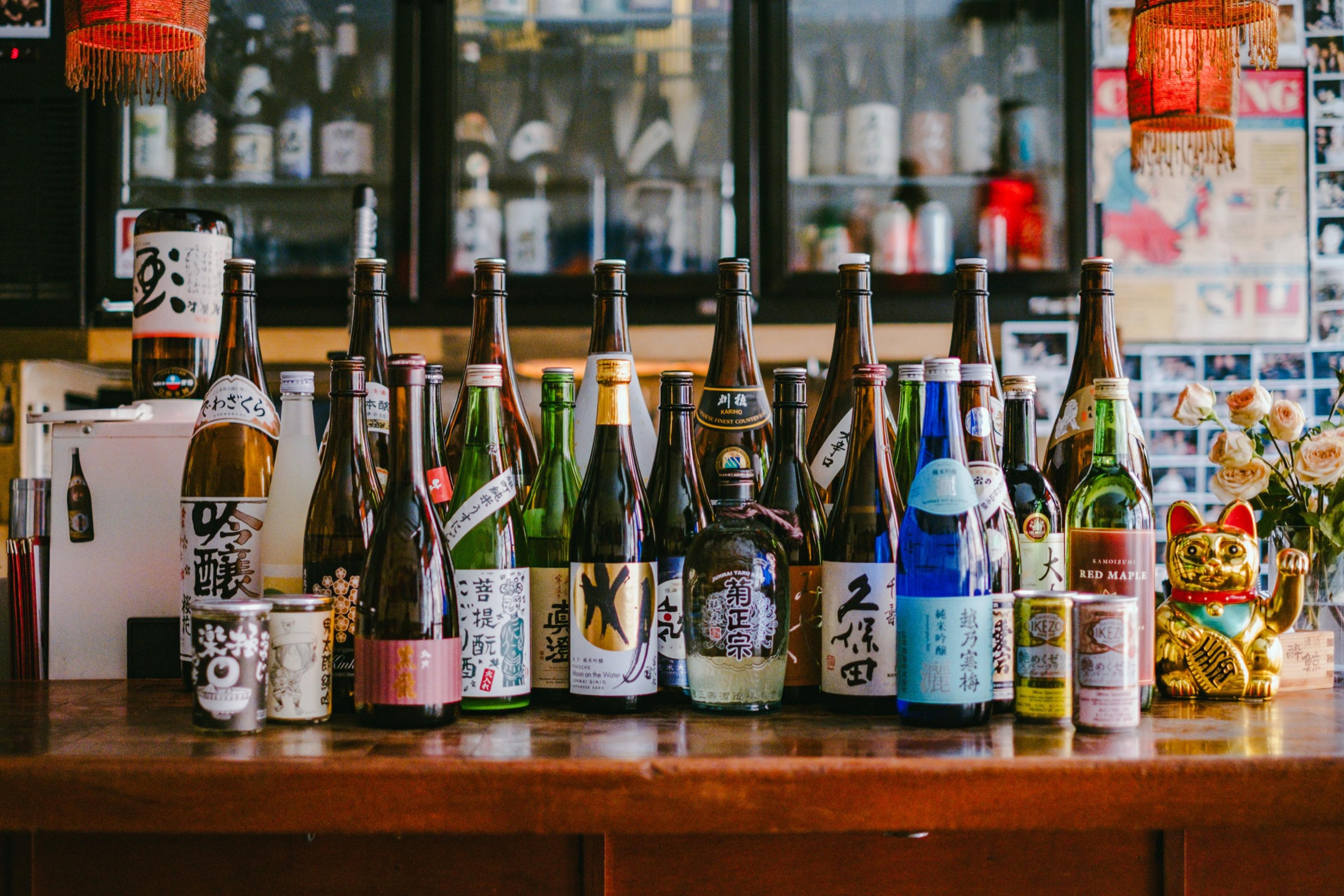 Rows of sake bottles on shelves behind a small bar, paper lanterns above