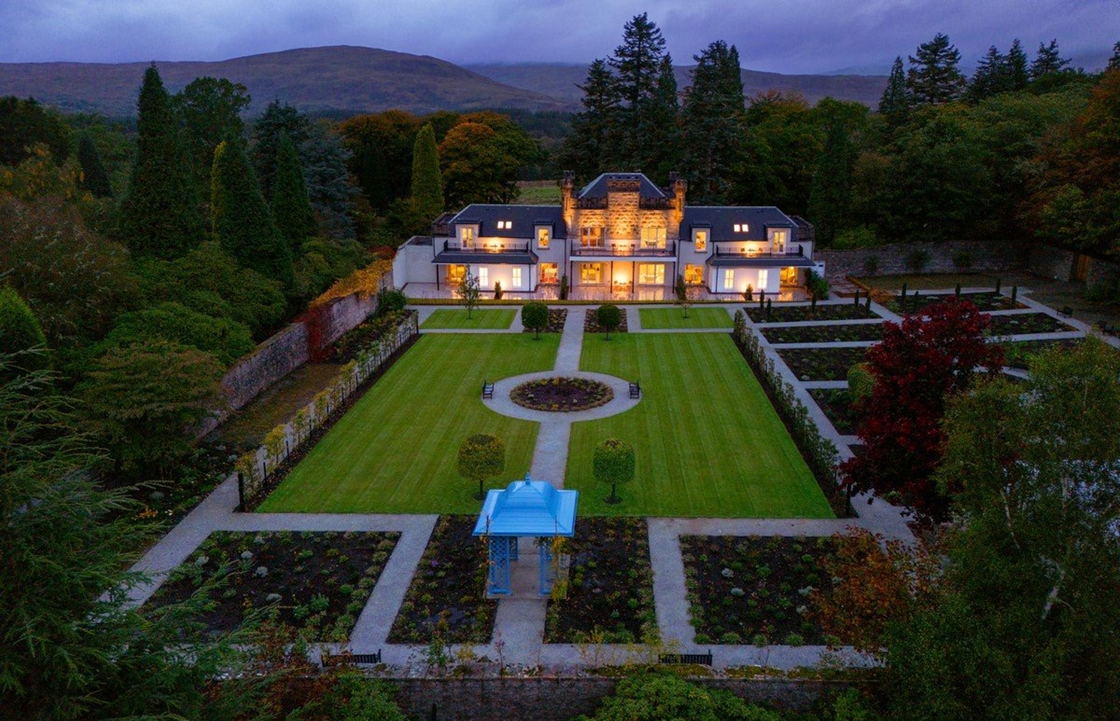 An aerial view at dusk of a white two-storey house with lit windows, set behind a formal walled garden with clipped box hedges.
