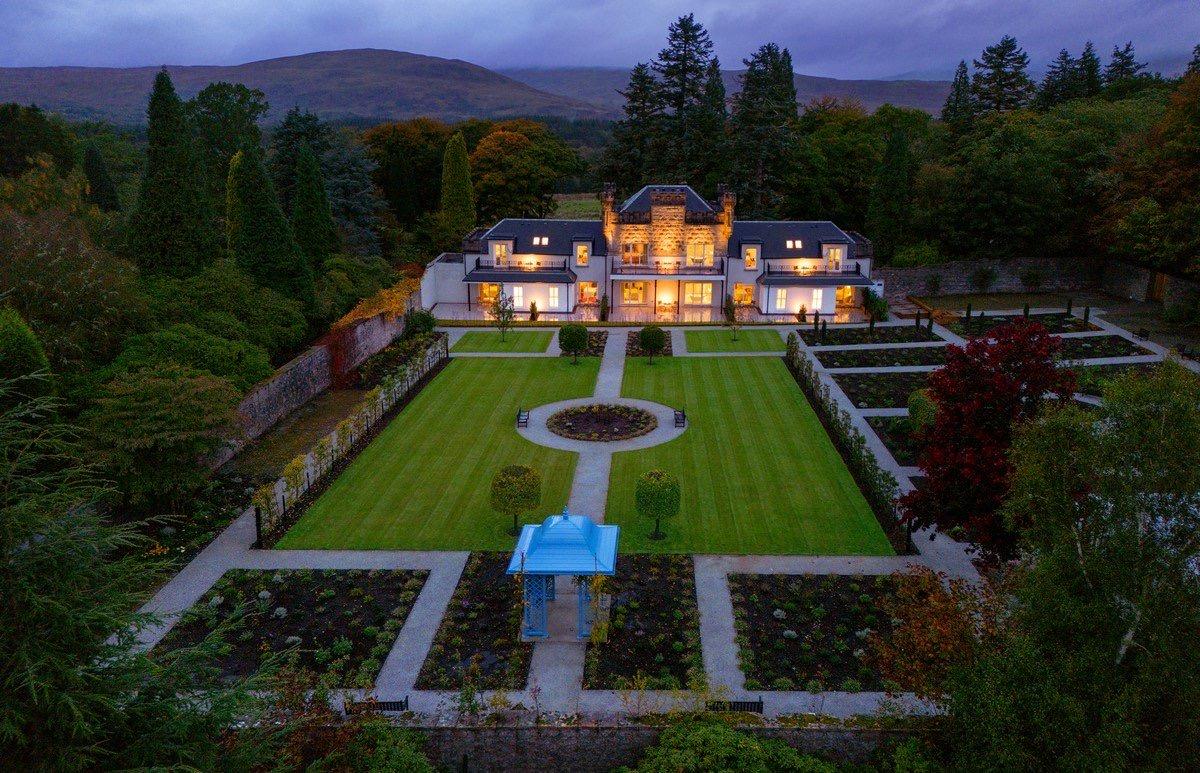 An aerial view at dusk of a white two-storey house with lit windows, set behind a formal walled garden with clipped box hedges.
