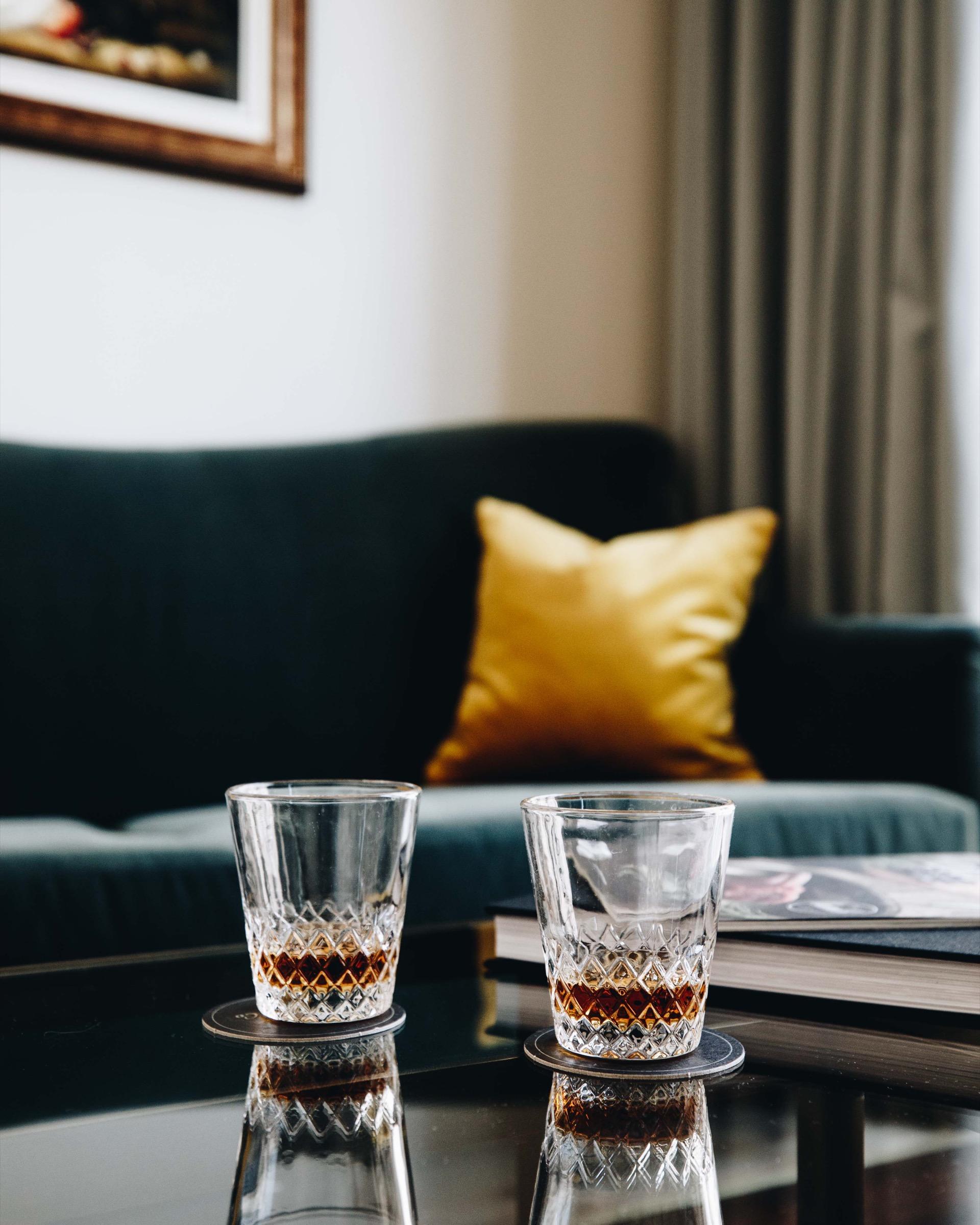 Two glasses of whisky on coasters on a glass table, with a book and a mustard cushion on a dark green sofa behind.