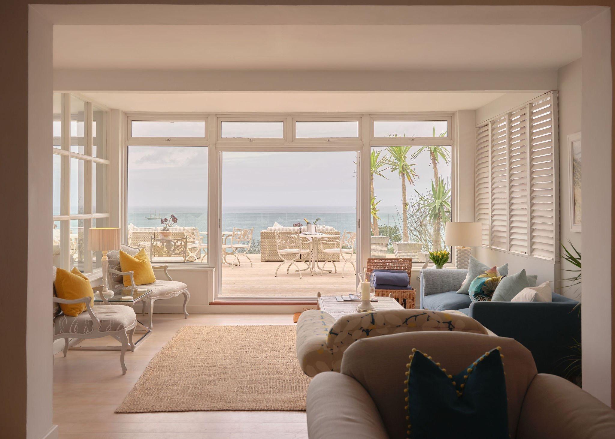 A living room with blue and cream sofas opening through large windows onto a terrace with dining furniture and the sea beyond.
