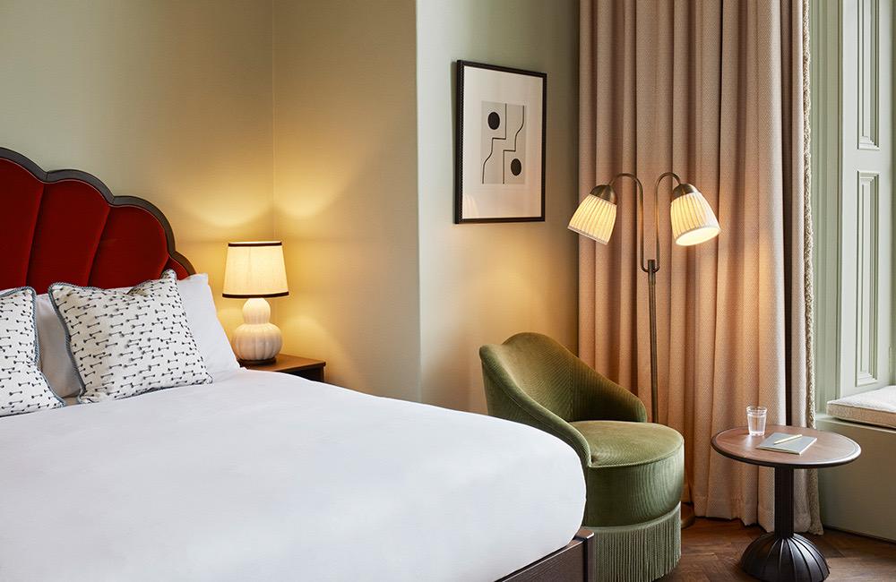 A bed beside a window hung with pink curtains, a green velvet armchair and a standing lamp in the corner.