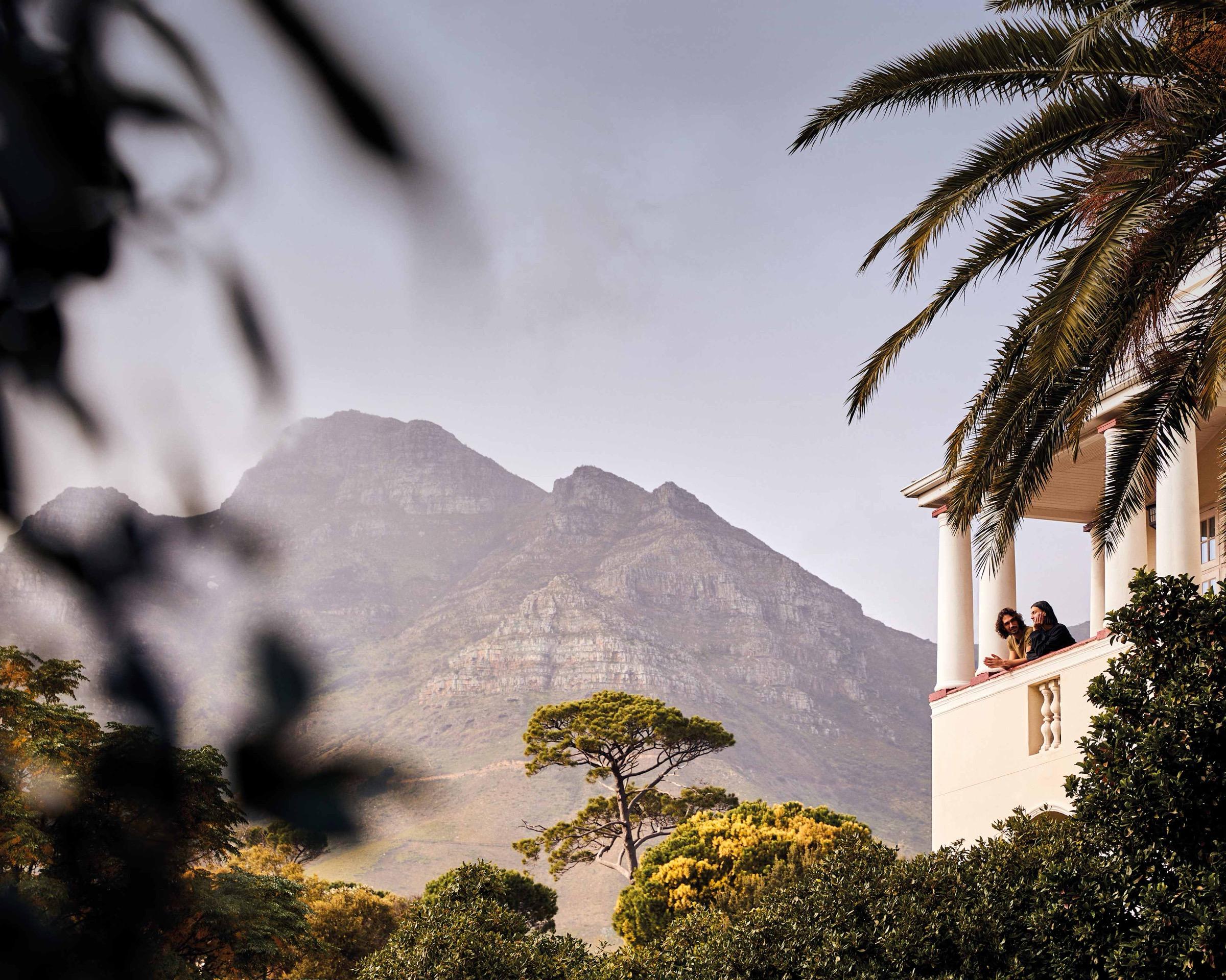 Two people lean on a white balcony with columns, looking out over trees towards a mountain ridge and a palm tree.