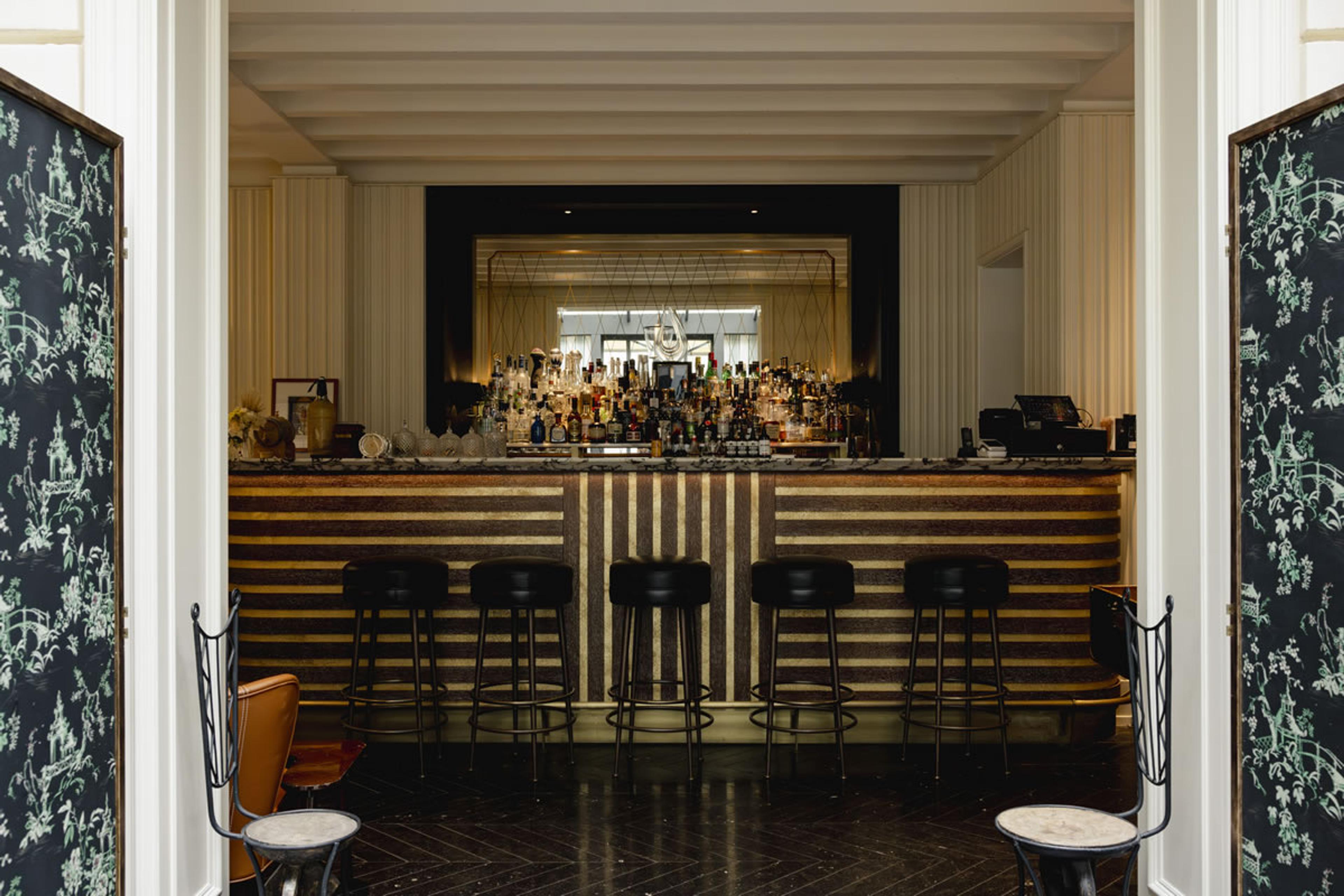A bar seen through a pair of patterned double doors, with a striped front, high stools and a mirrored shelf of bottles.