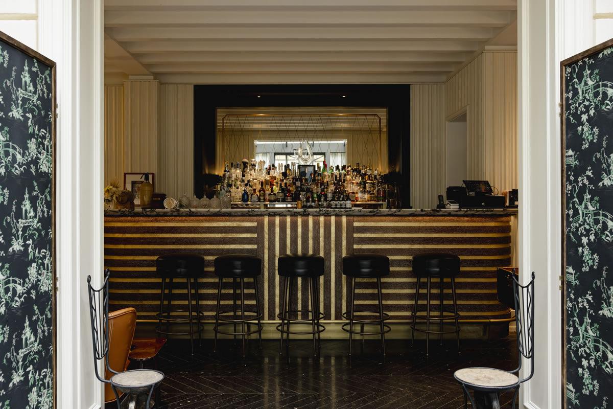 A bar seen through a pair of patterned double doors, with a striped front, high stools and a mirrored shelf of bottles.