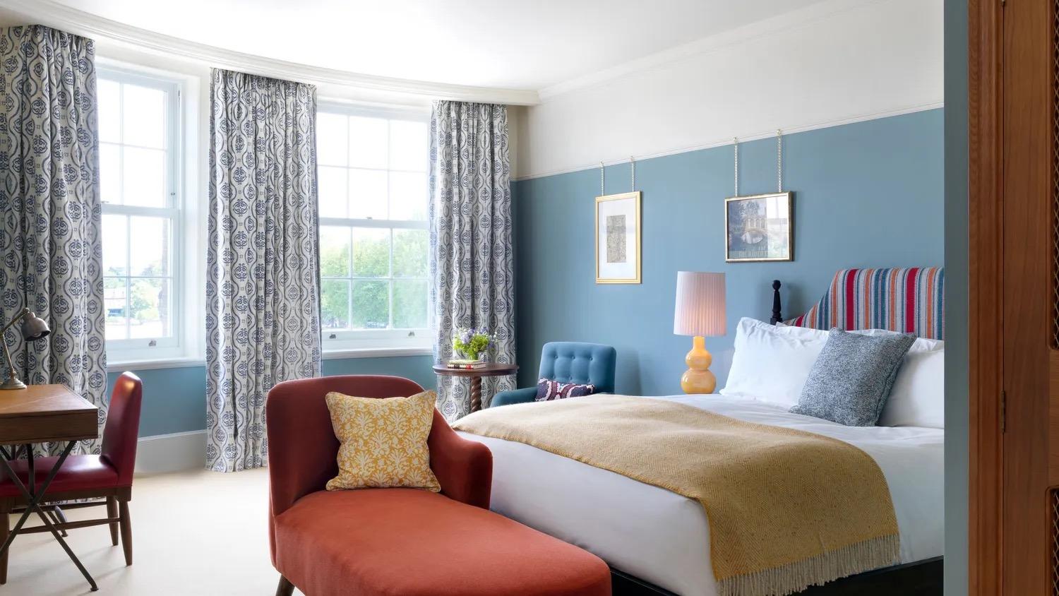 a bedroom with blue walls, a bed with a striped headboard, a rust coloured chaise longue at its foot, and two curtained windows