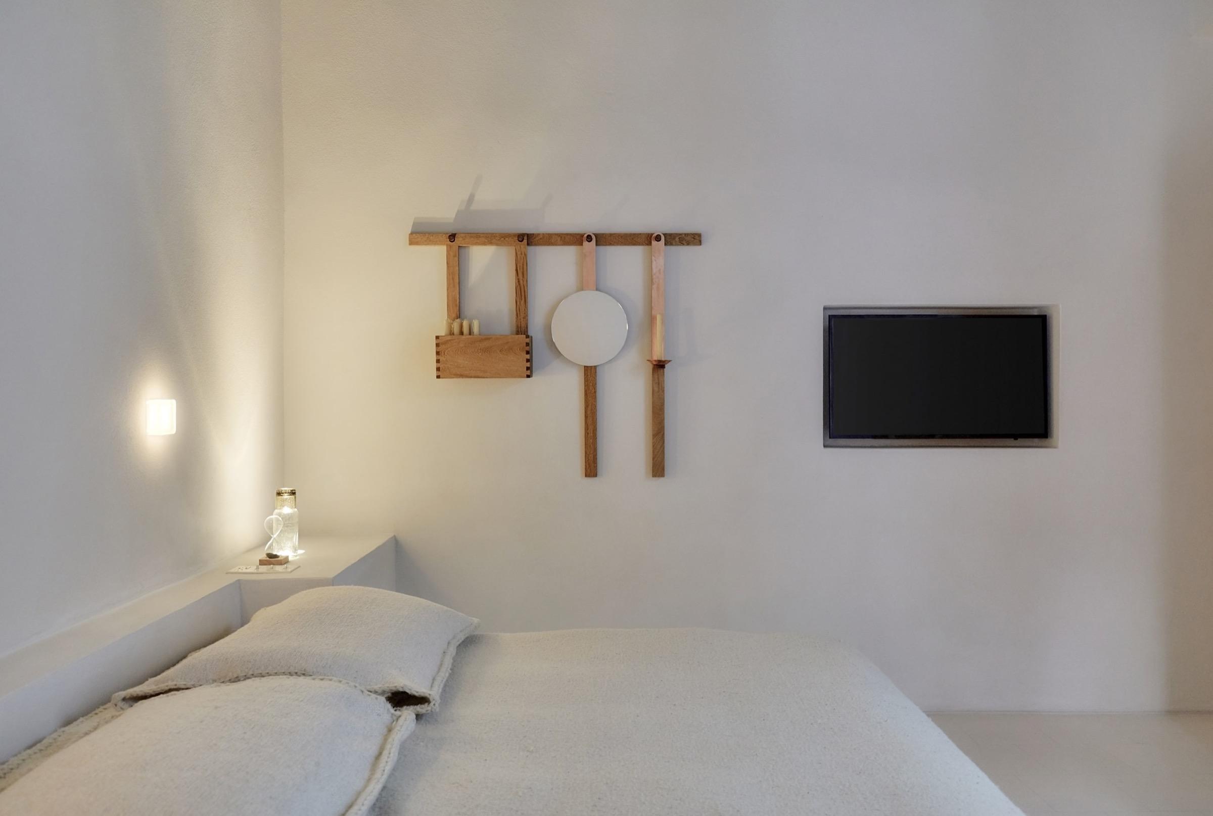 A bed with white linen and pillows, a wooden wall rack above the headboard, a mounted television, and a bedside lamp.