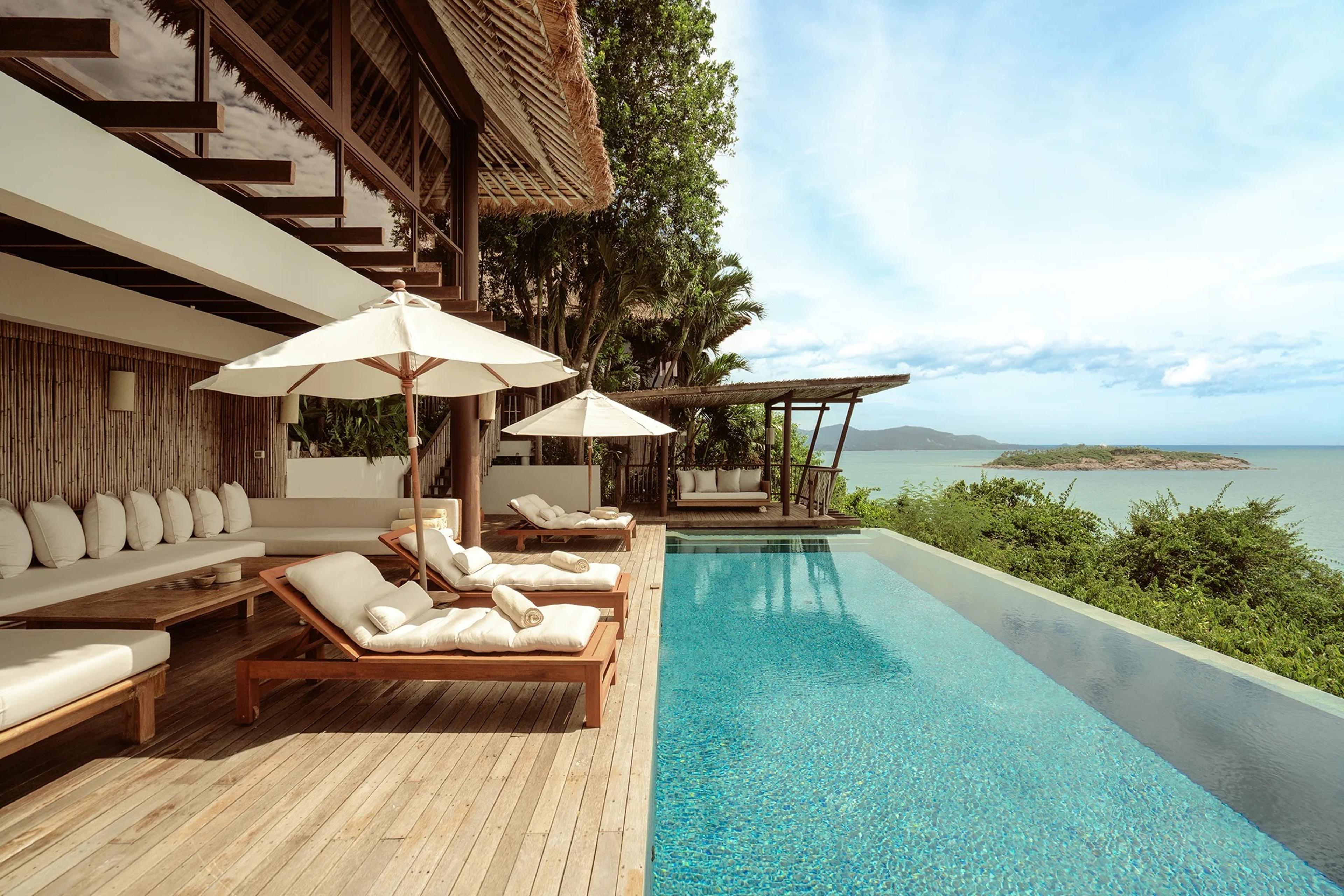A wooden pool deck with cushioned loungers and umbrellas runs along a thatched-roof building, beside an infinity pool looking out over the sea and a small island.