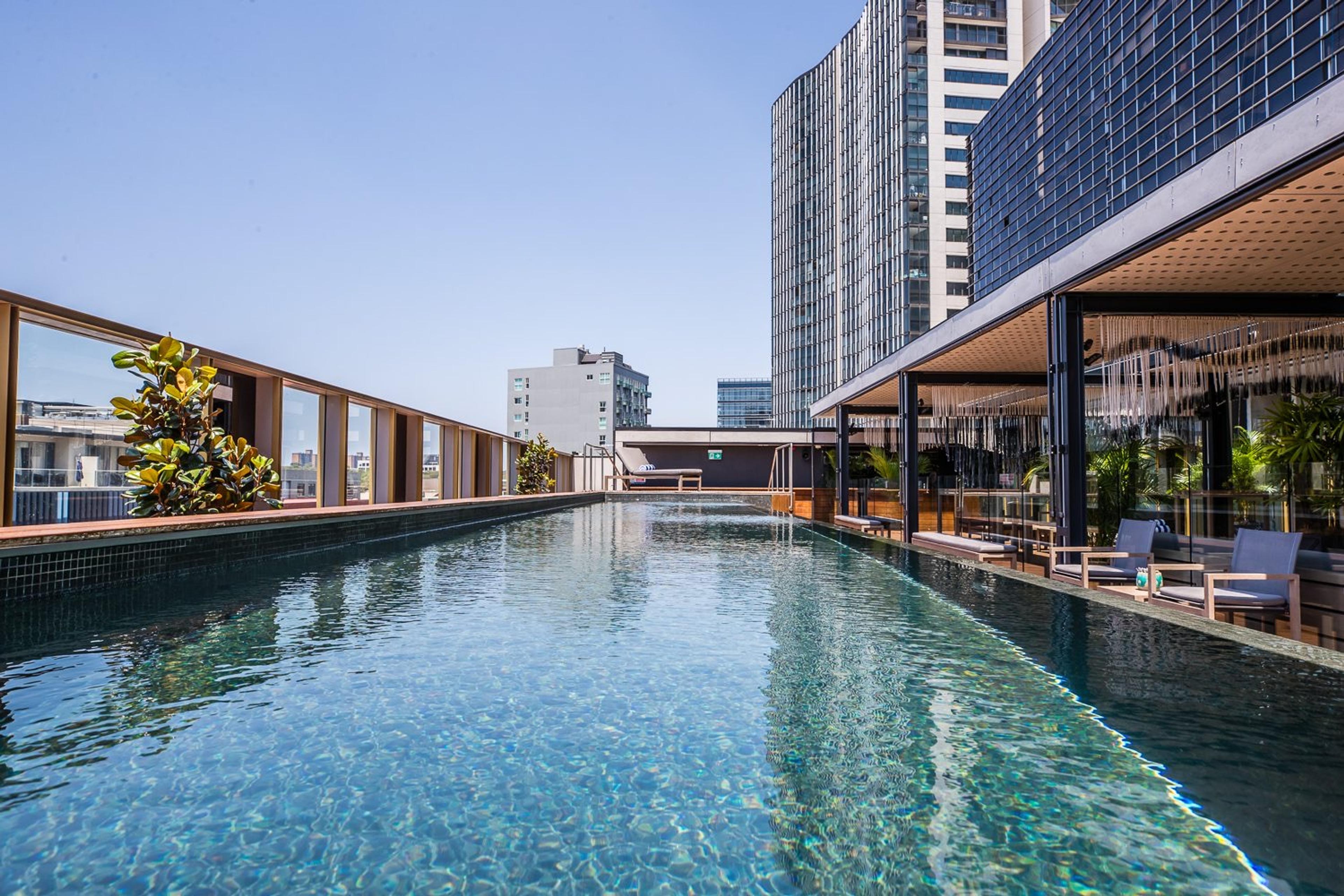 A rooftop lap pool beside a glass-walled lounge area, with sun loungers, plants, and high-rise towers against a clear sky.