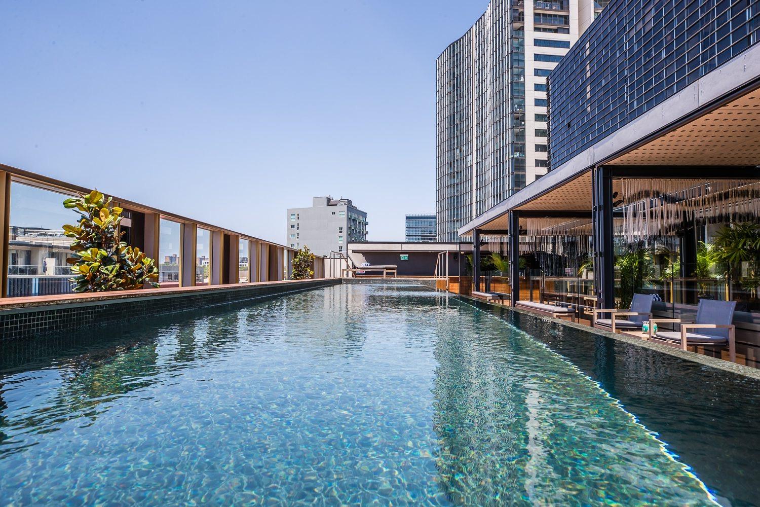 A rooftop lap pool beside a glass-walled lounge area, with sun loungers, plants, and high-rise towers against a clear sky.