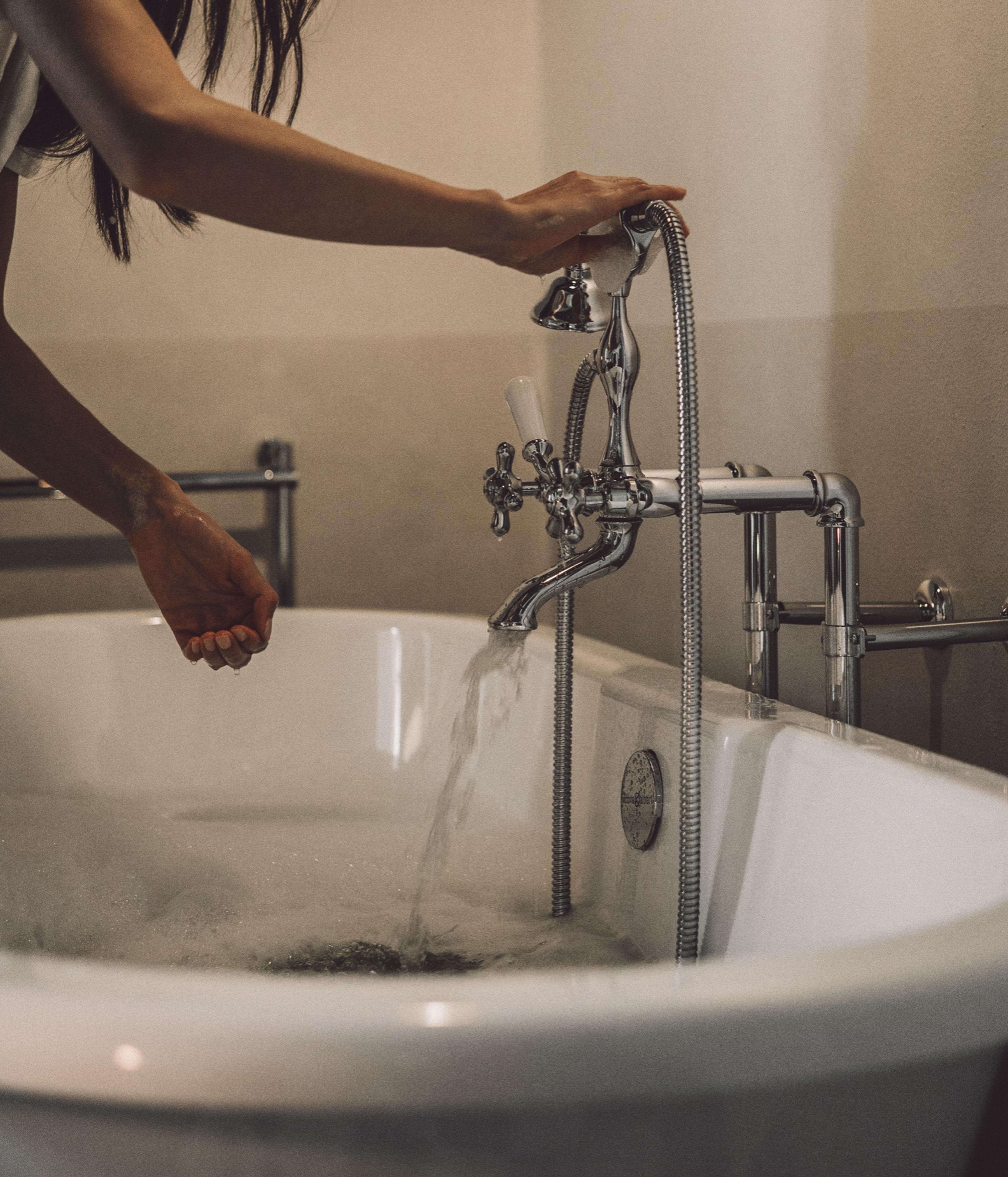 A hand holds a shower attachment over a freestanding tub filling with water, chrome pipework and taps beside it.