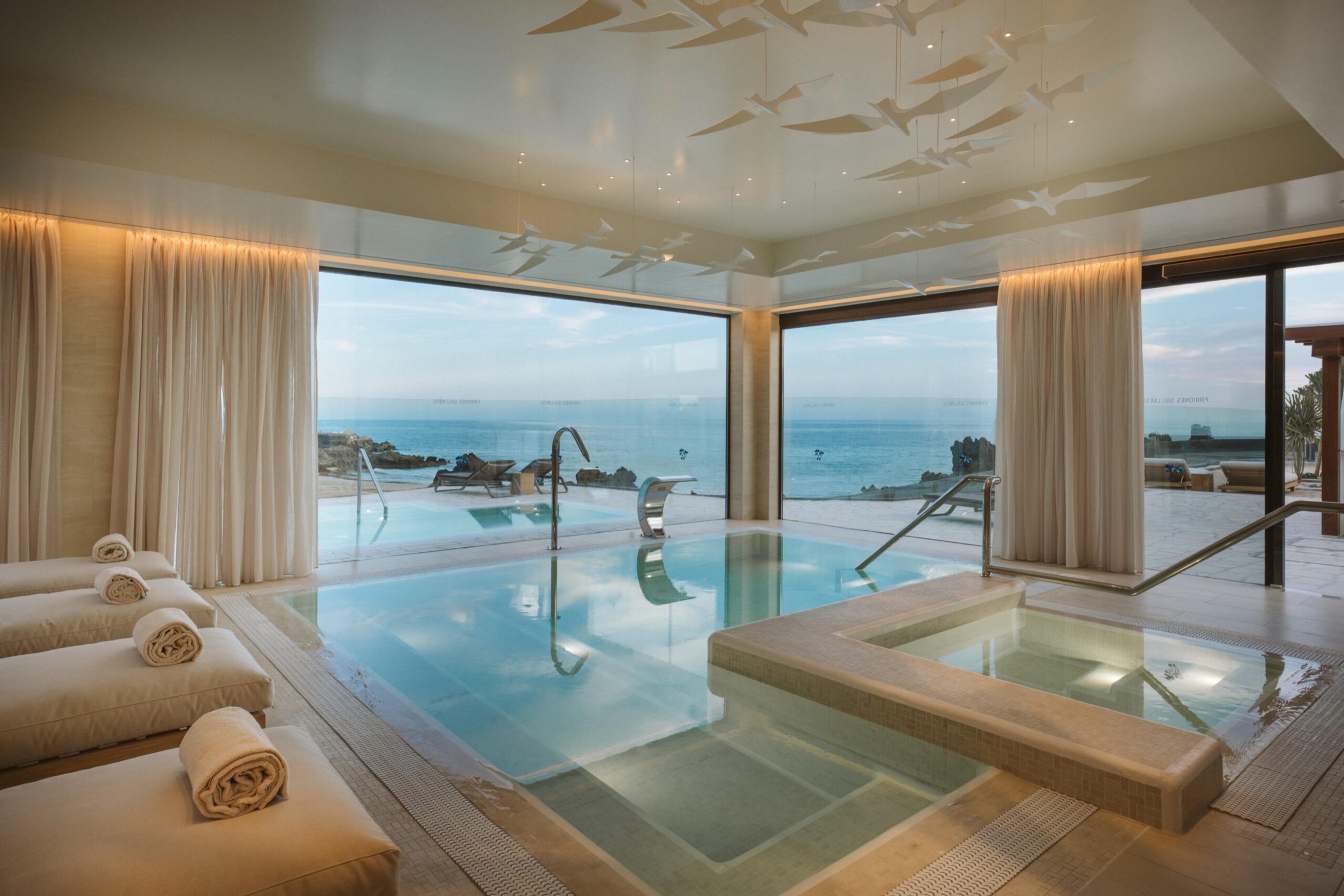 An indoor spa pool and square hot tub with a stepped edge, rows of folded towels on a bench in the foreground and glass doors open to loungers, rocks and the sea.