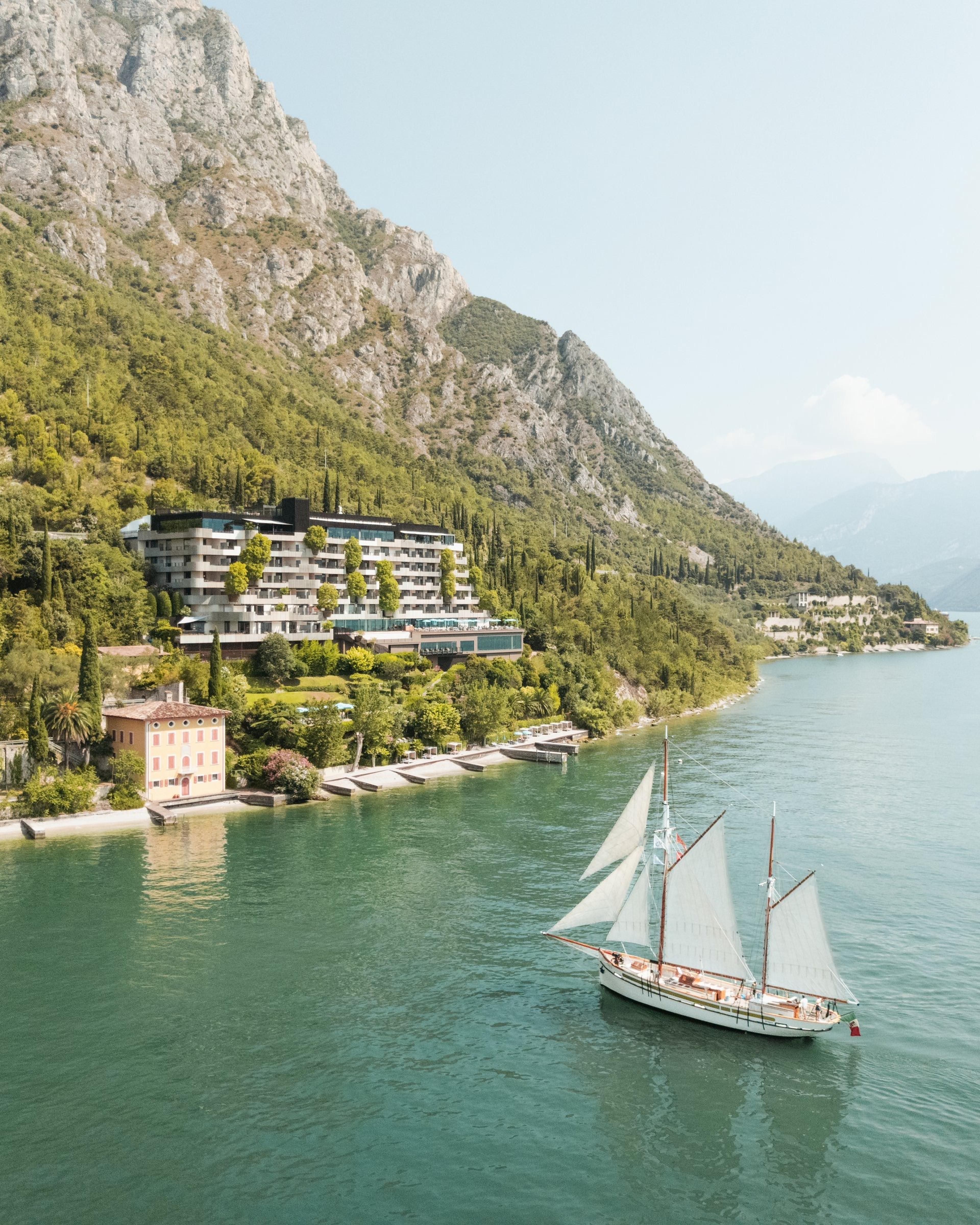 an aerial view of a terraced hotel building on a wooded lakeshore, with a sailing boat passing in the foreground