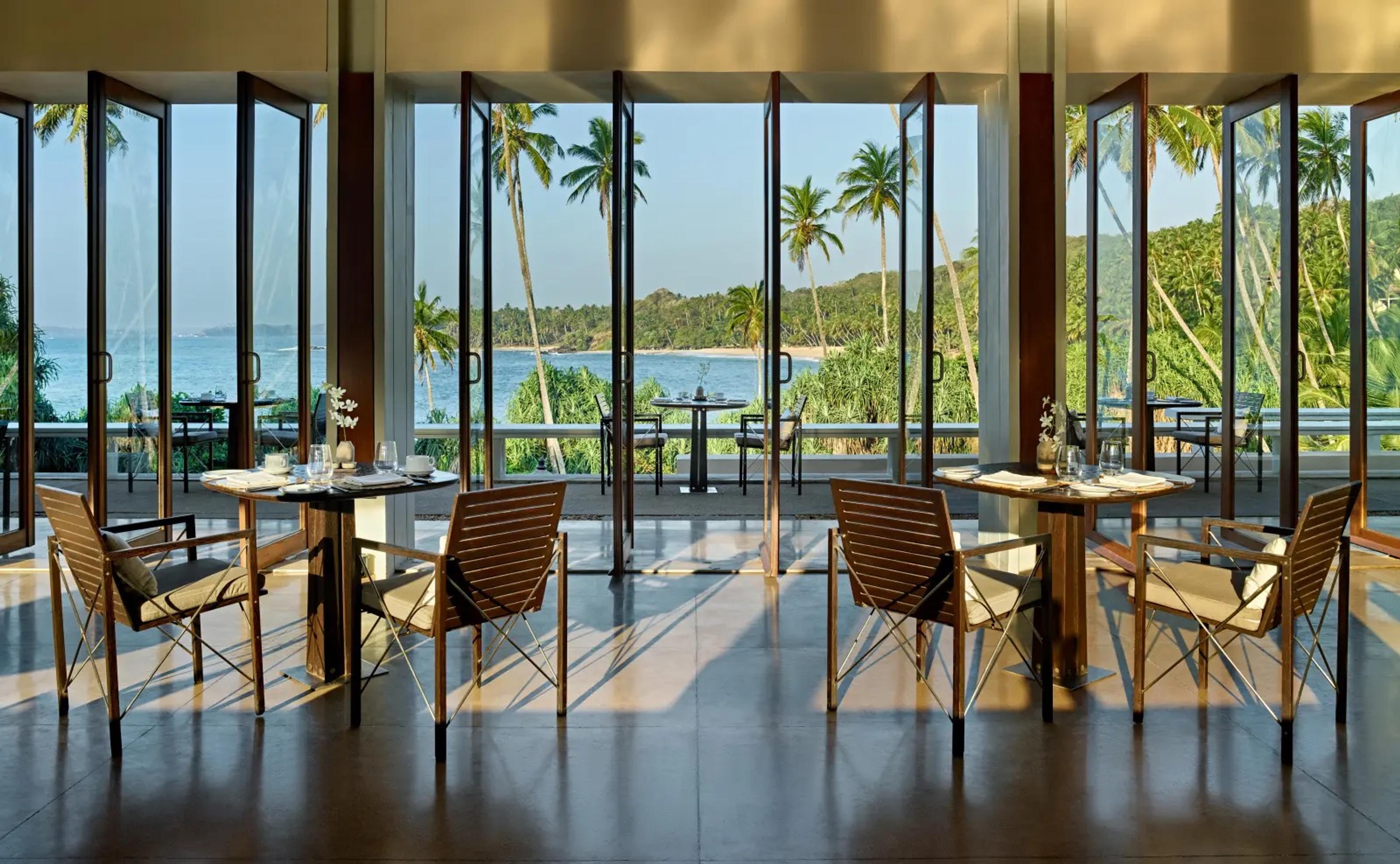 A dining room with floor to ceiling doors open on both sides, tables set for lunch and a view across a terrace to the sea and palm trees.