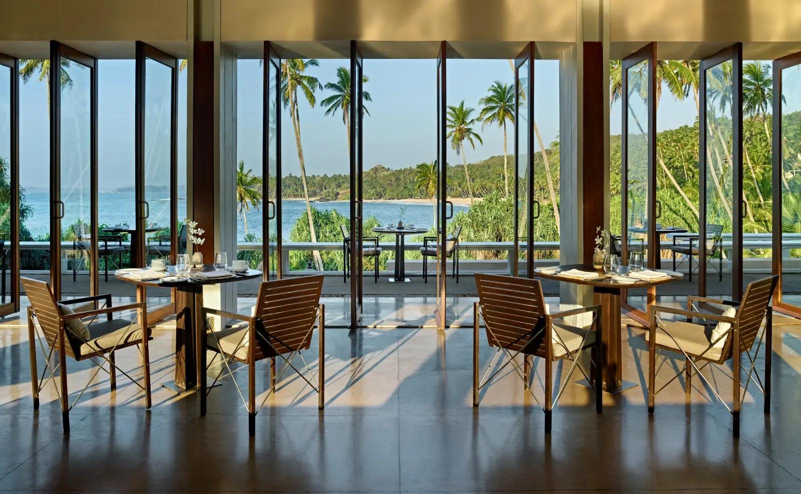 A dining room with floor to ceiling doors open on both sides, tables set for lunch and a view across a terrace to the sea and palm trees.