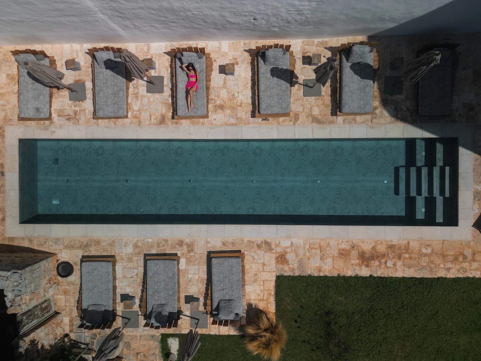 An overhead view of a long rectangular swimming pool and a row of sunbeds, with one woman lying on a lounger.