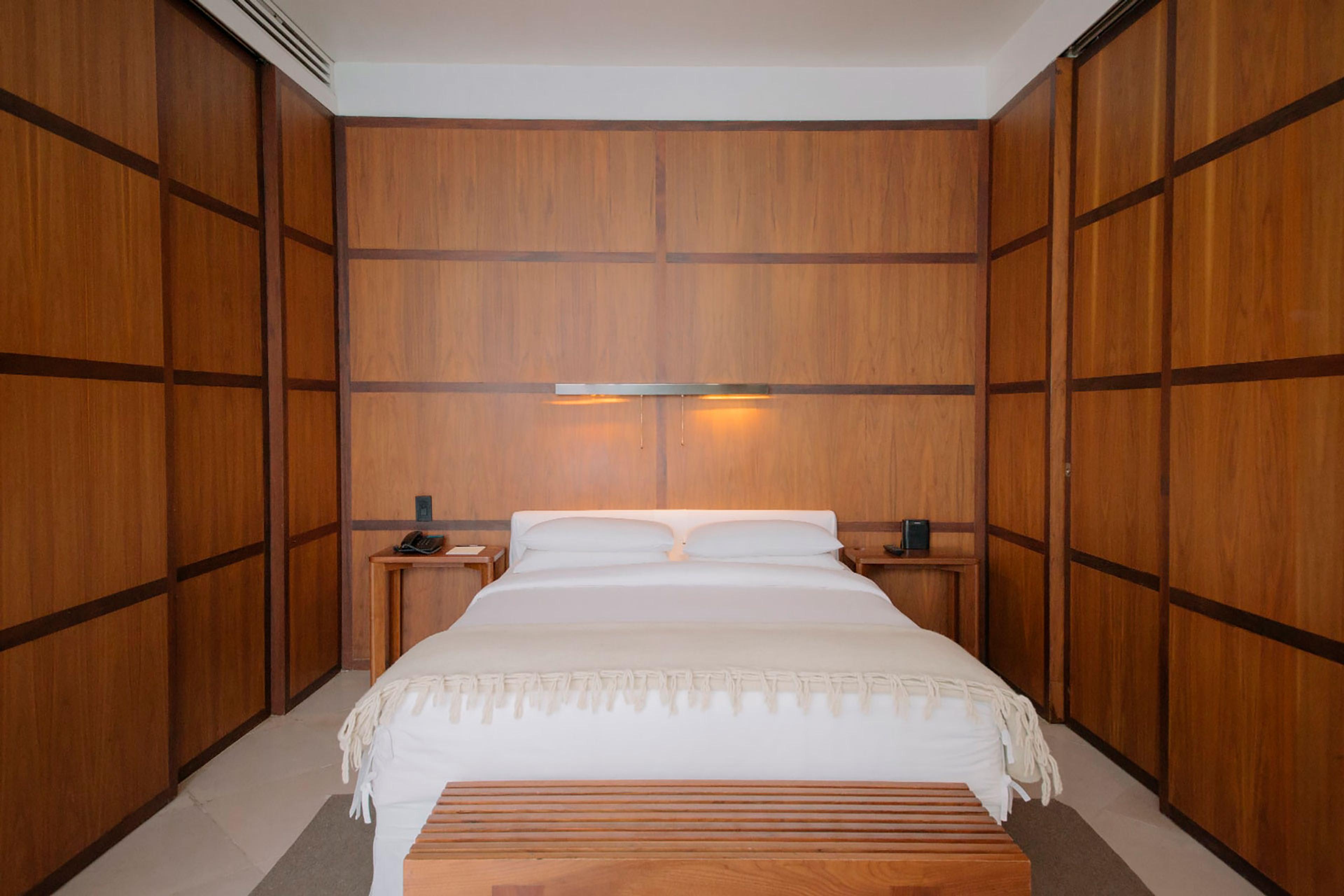 A bedroom fully panelled in dark wood, with a white bed centred between matching nightstands and a wooden bench at its foot.