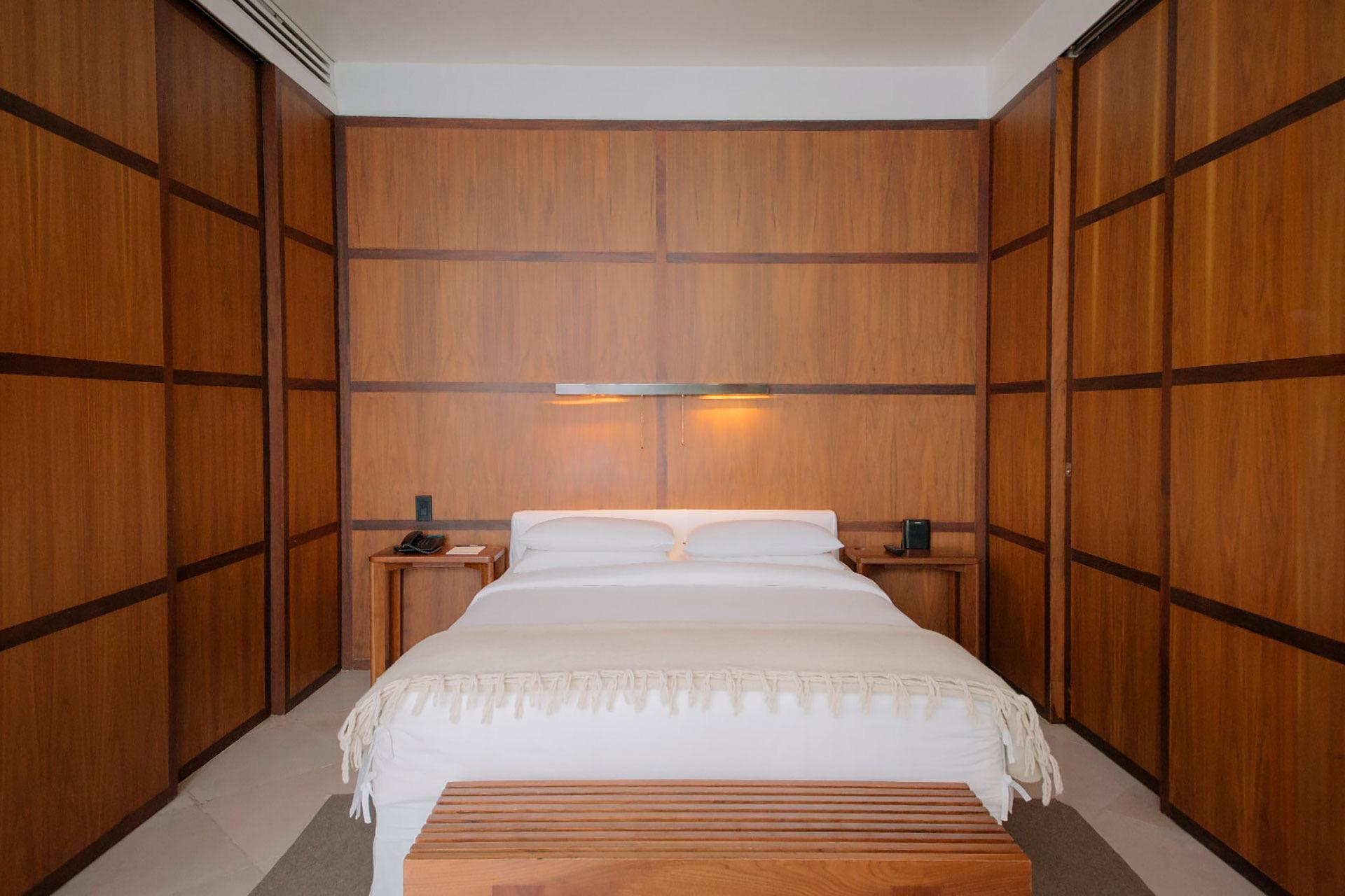 A bedroom fully panelled in dark wood, with a white bed centred between matching nightstands and a wooden bench at its foot.