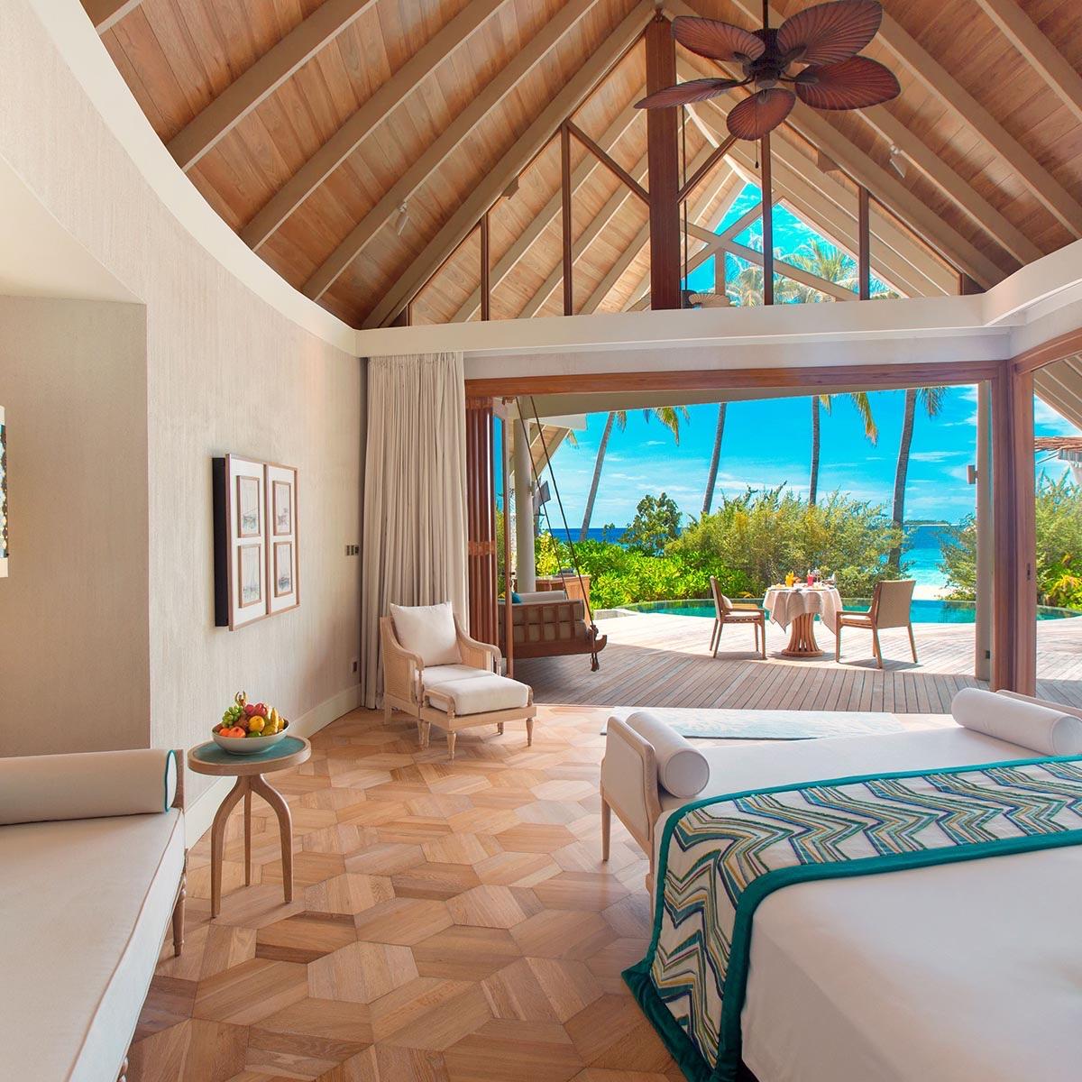 An open-sided bedroom under a high wood-raftered roof, with a bed and a daybed facing a deck set for dining beside a pool and trees.