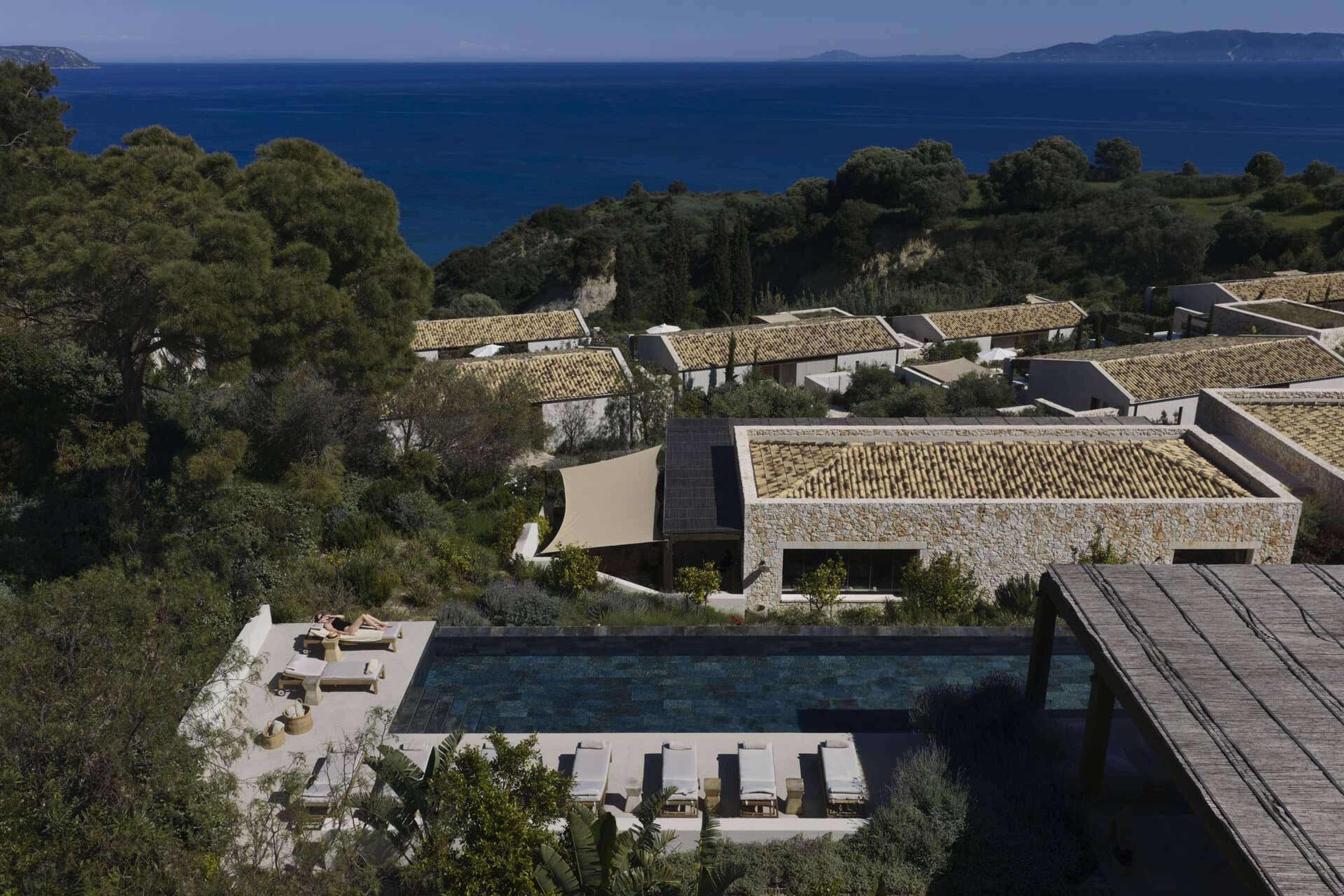 an elevated view of the hotel's stone and white buildings and terracotta roofs around a pool, with one person on a lounger, trees and the sea beyond