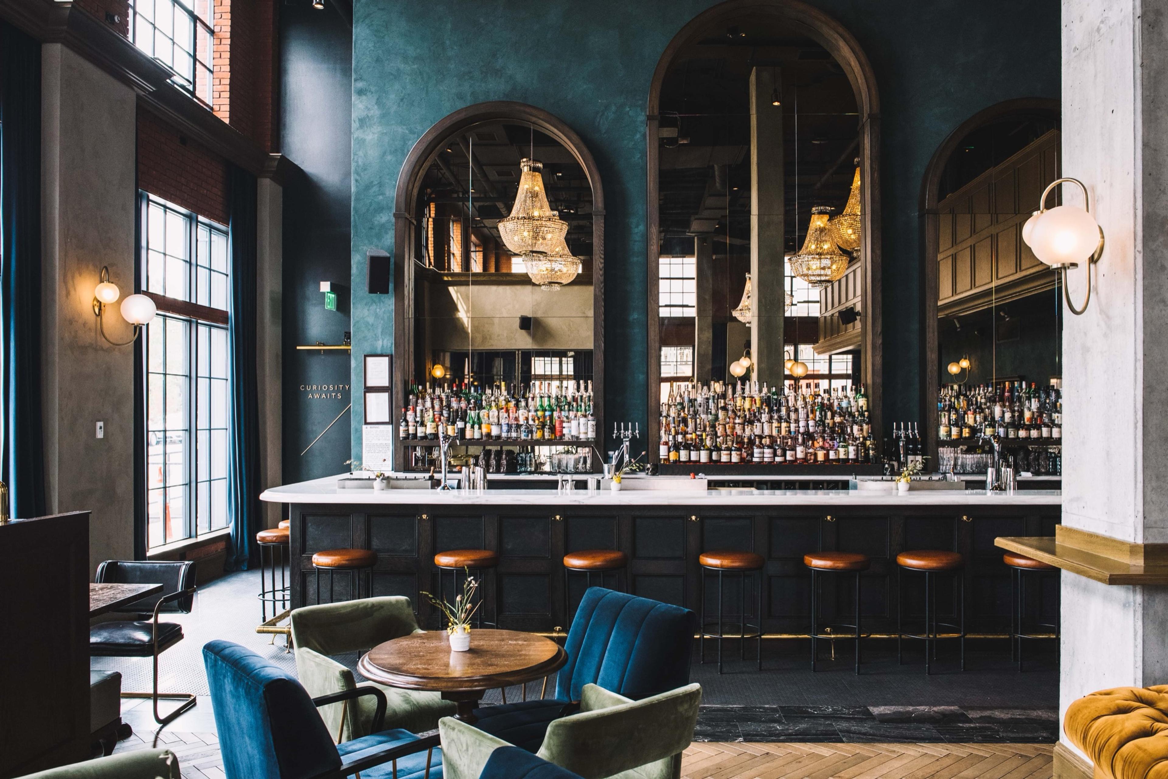 A hotel bar beneath three arched mirrors and chandeliers, seen across a sitting area of blue velvet armchairs around a round table.