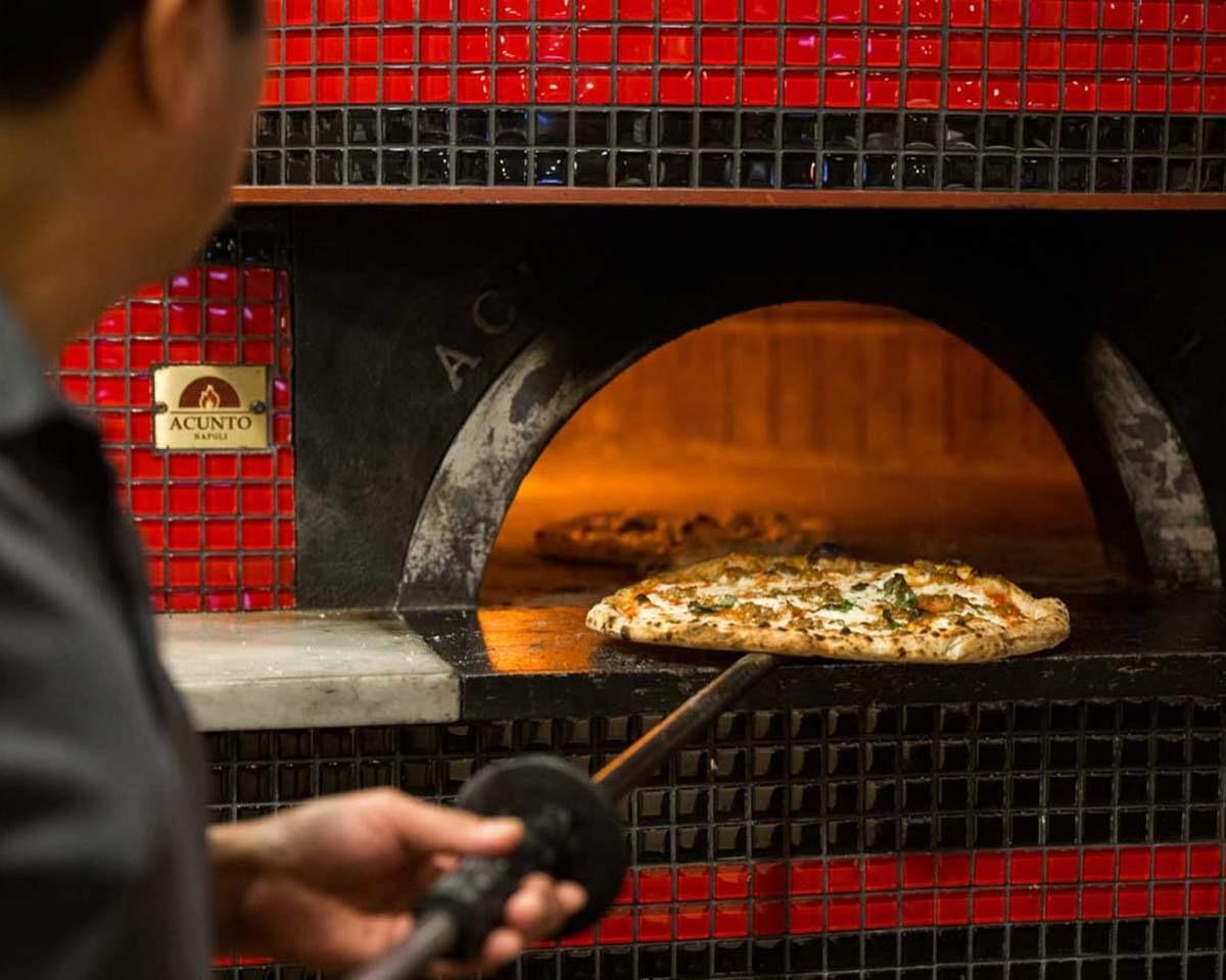 A pizza on the peel at the mouth of a wood-fired oven tiled in red