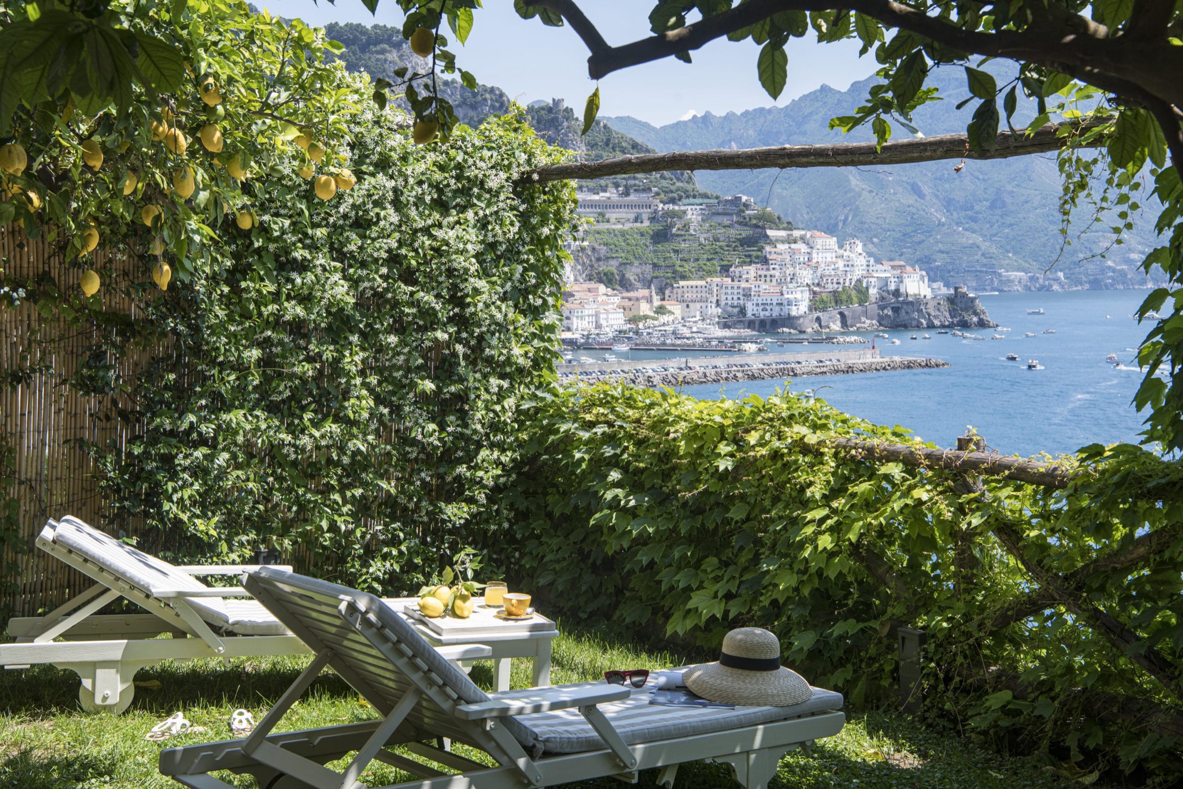 Two sun loungers under a lemon tree, with a breakfast tray, sunglasses and a straw hat, looking over a hillside town and the sea.
