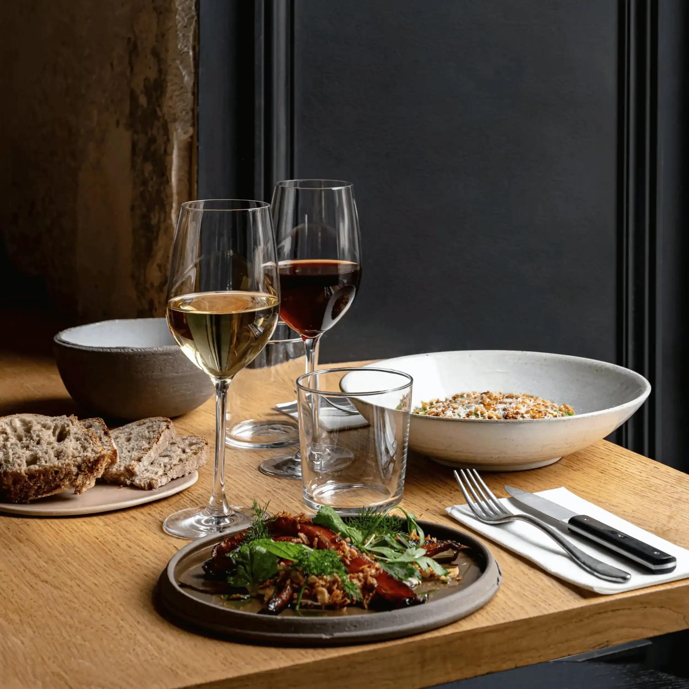 A wooden restaurant table set with two glasses of wine, a bowl of grain salad, a plate of char-grilled vegetables with herbs, and torn bread.