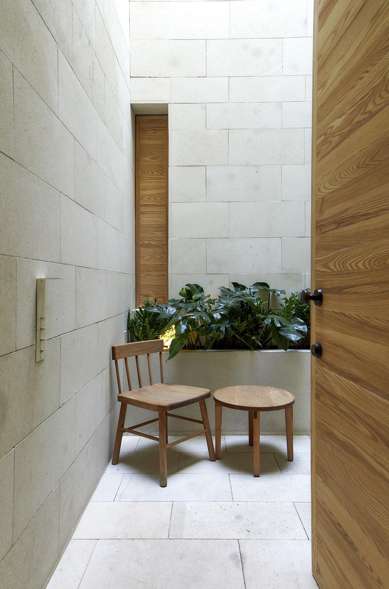A small stone-walled nook with an open wooden door, a single wooden chair and round side table, and potted plants by a window.