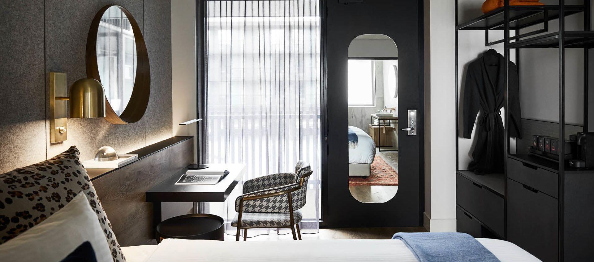 a hotel room with a made bed, a desk and chair by the window, and an open black door reflecting the bathroom, with a robe hanging in the closet