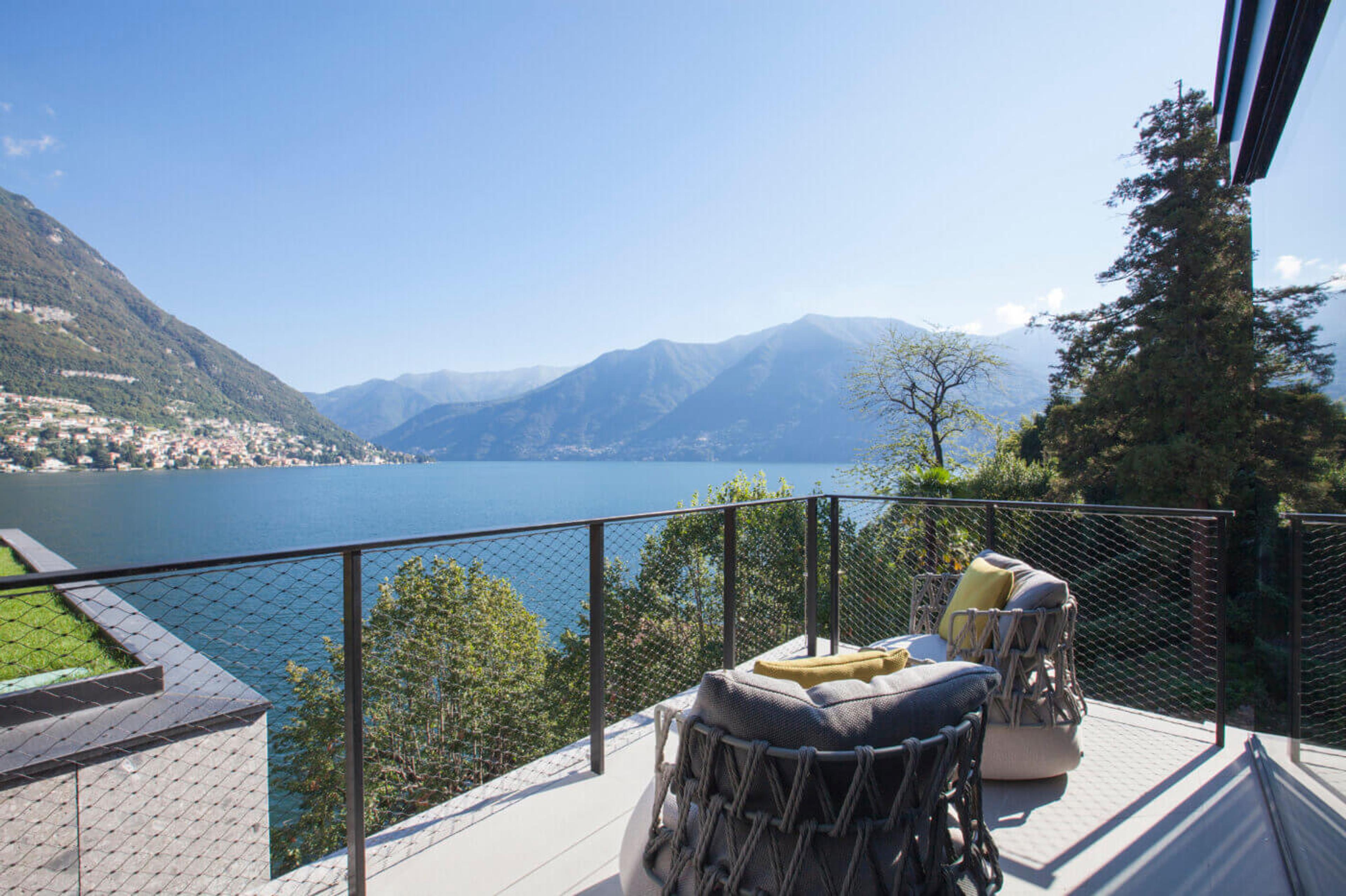 A wide view over a lake and its surrounding mountains from a terrace, with the edge of a building visible at the top of the frame.