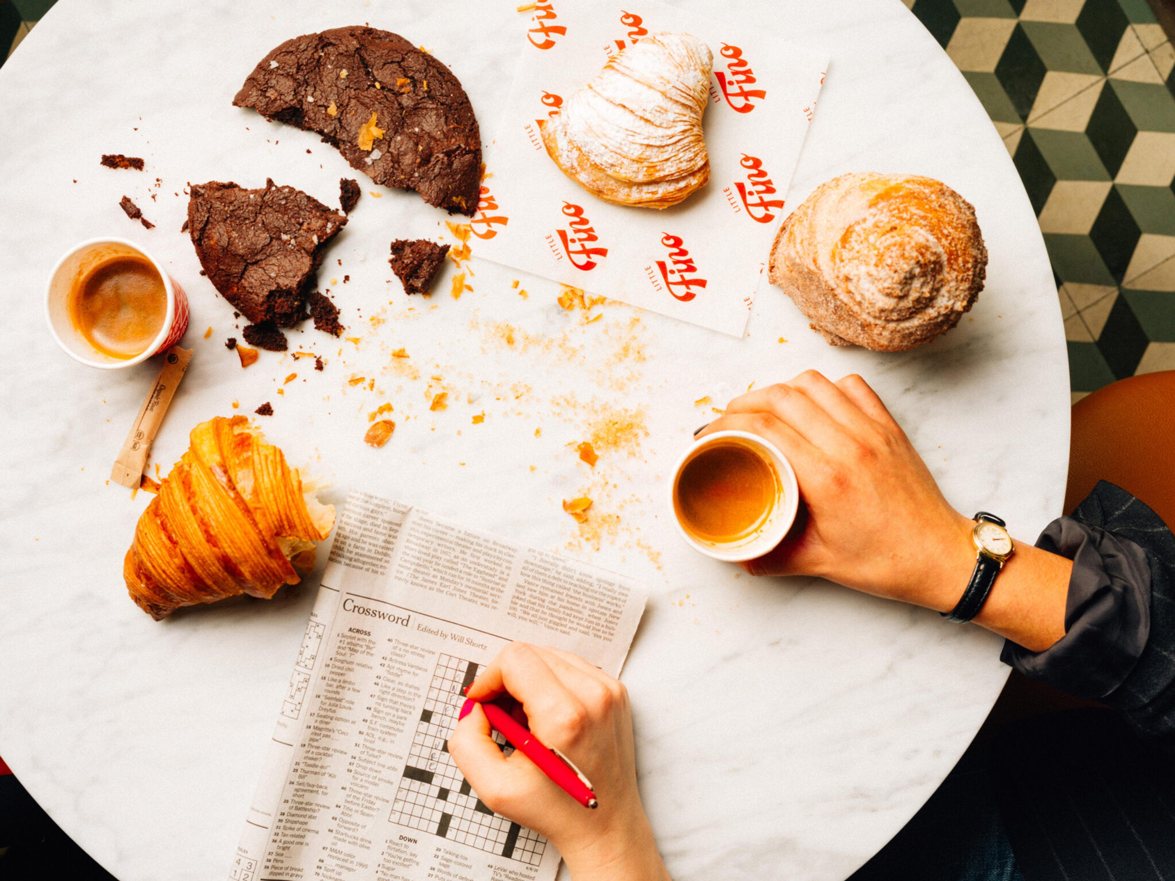 A marble cafe table scattered with pastry crumbs, two cups of coffee, a half-eaten cookie, a croissant, and a hand filling in a newspaper crossword.