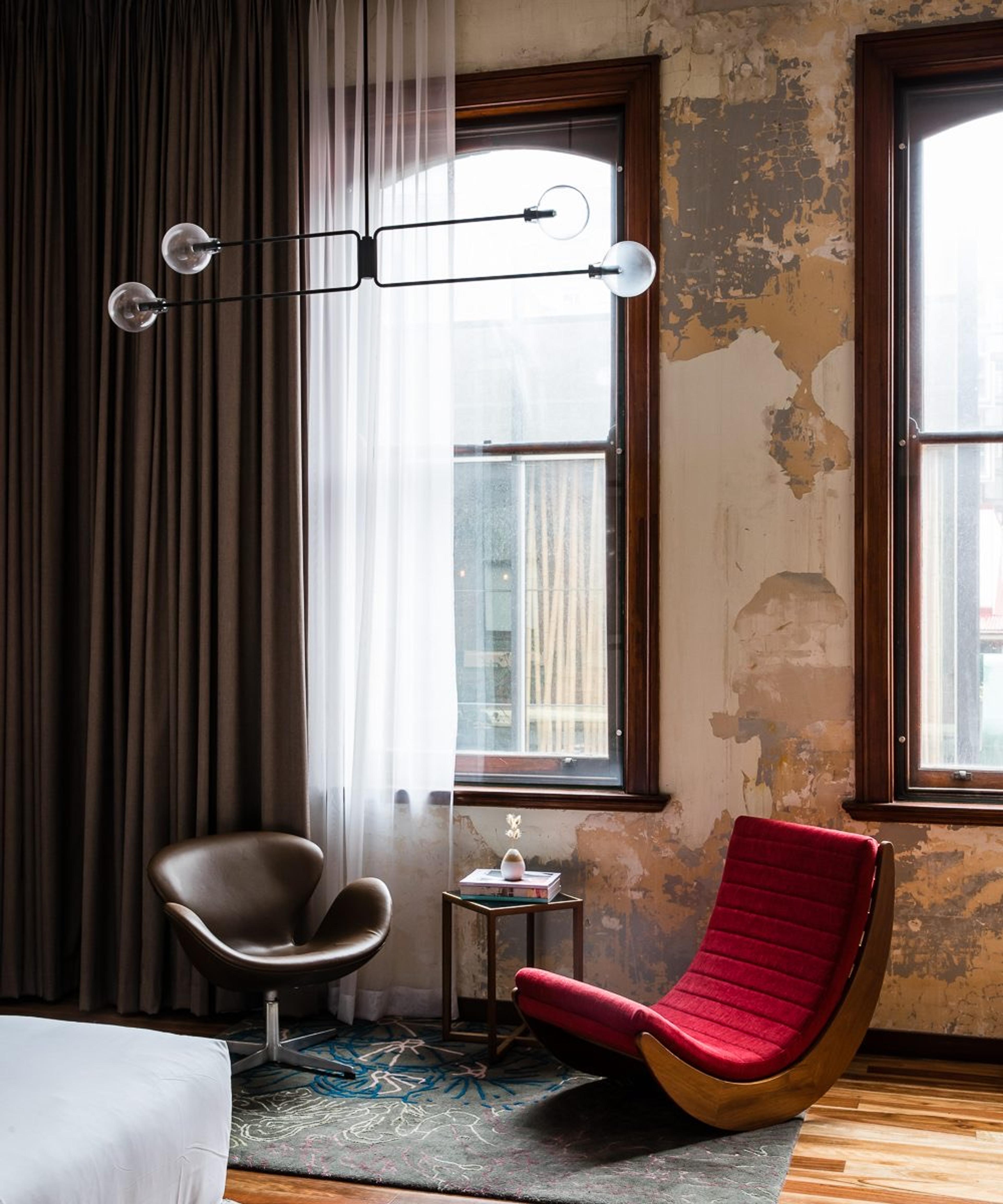 A room with a patched and peeling plaster wall, two tall arched sash windows, a brown swivel chair, and a red curved lounge chair on a patterned rug.