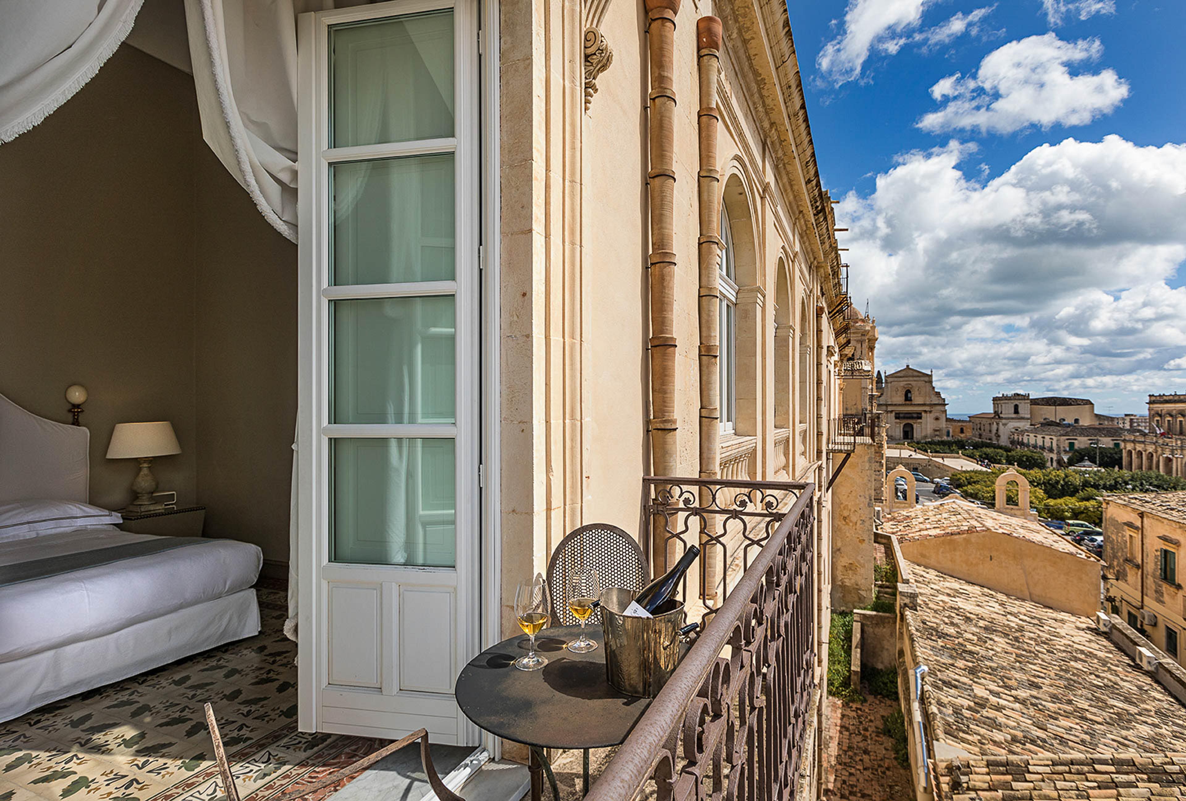 A hotel balcony table set with two glasses of white wine and a wine bucket, an iron railing overlooking terracotta rooftops and a domed church in Noto.