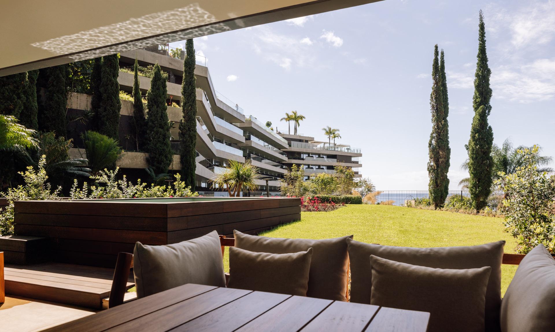 A shaded terrace with cushioned seating looking out over a lawn to the hotel's terraced building and the sea.