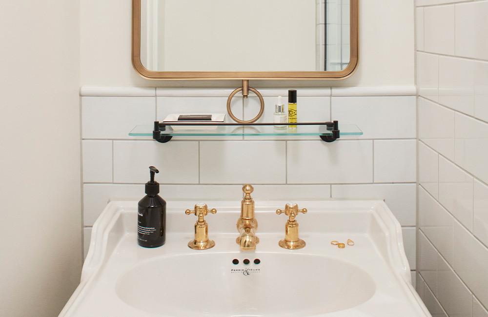 A white pedestal basin with brass taps under a round brass-framed mirror.