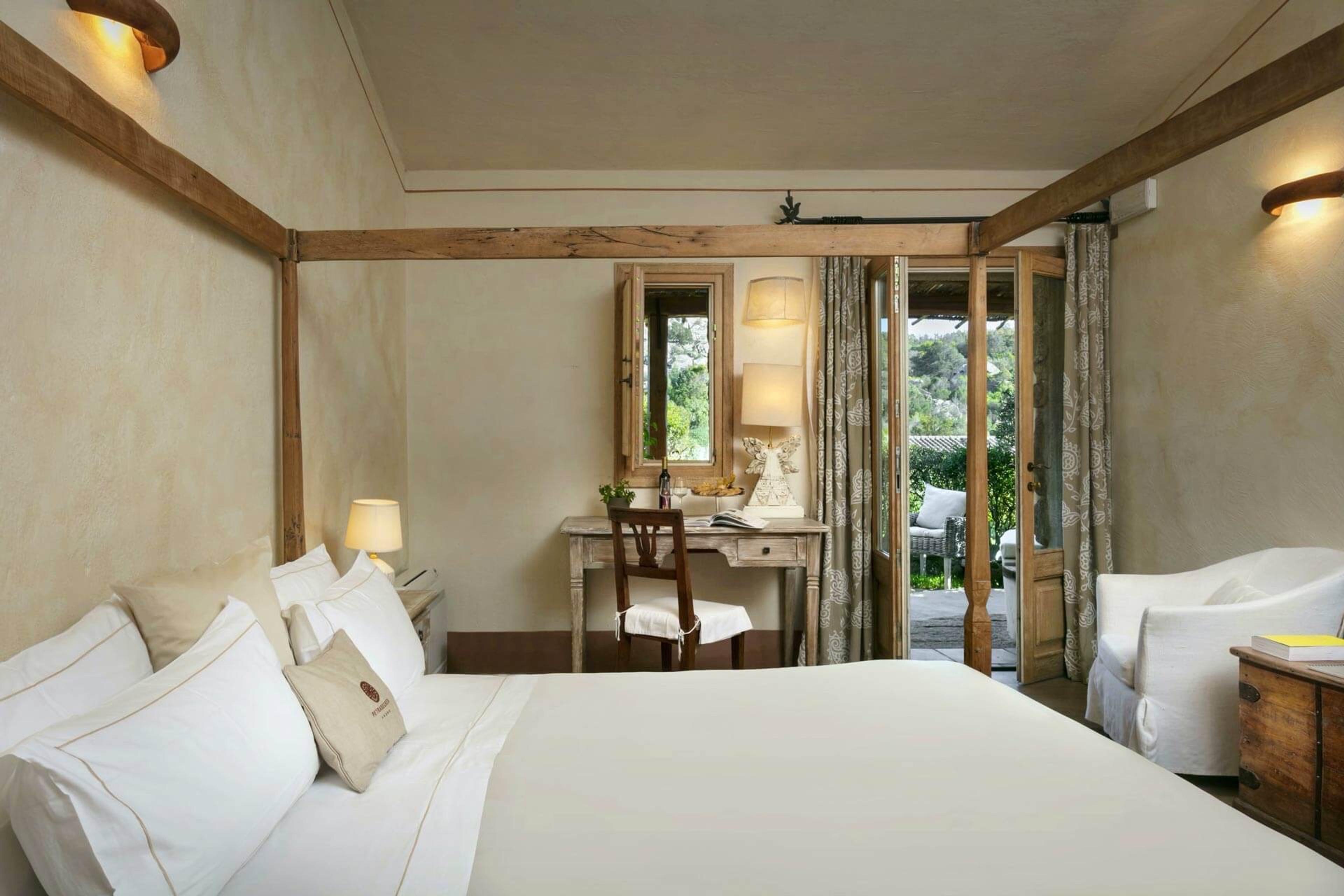 A bedroom with a wood-beamed ceiling, a white bed, a writing desk and an armchair, with glass doors open to a terrace.