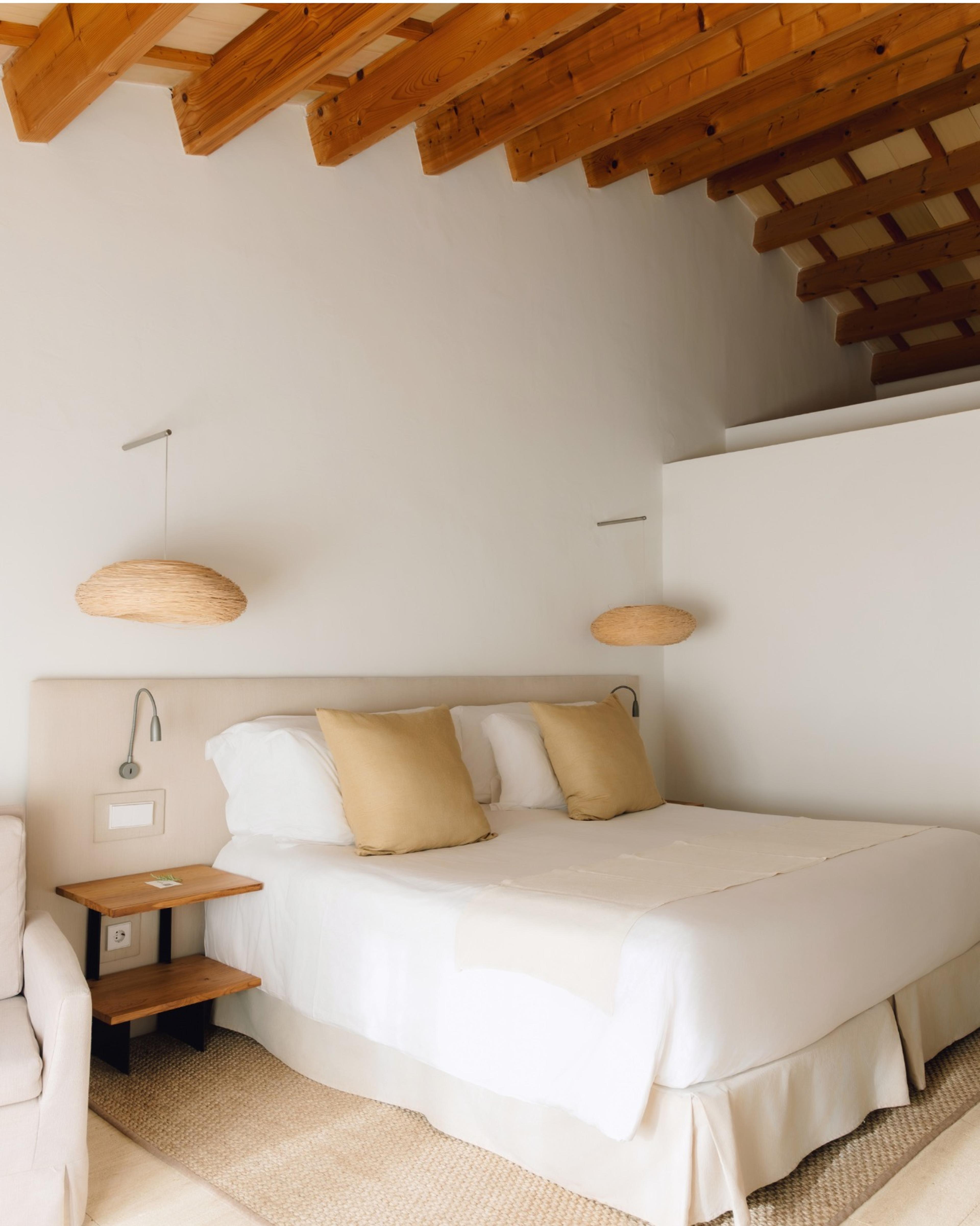 A neatly made double bed with cream linen and mustard cushions under an exposed timber ceiling, beside a small wooden nightstand.