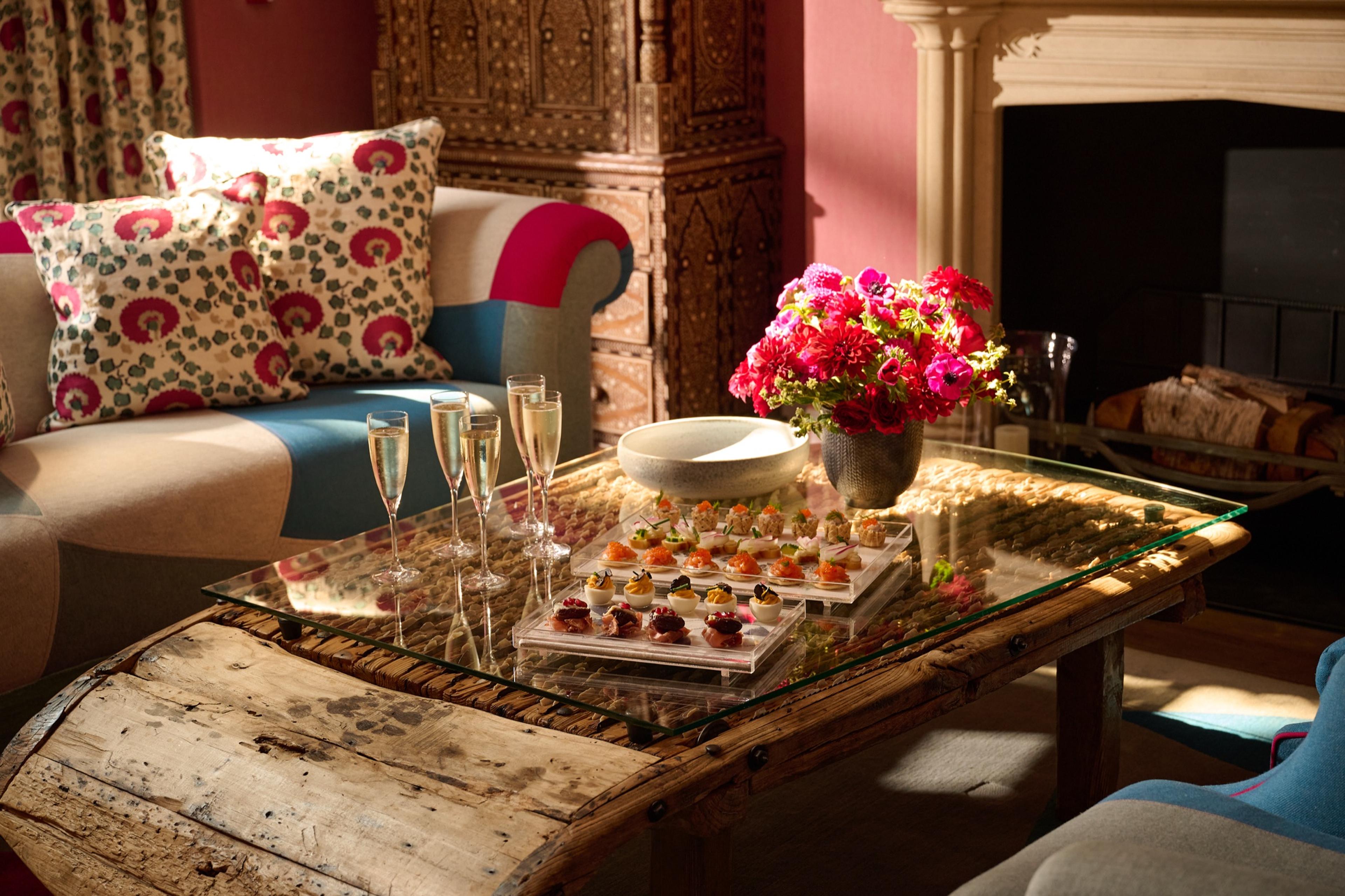 A coffee table with four glasses of champagne, a tray of canapes, a bowl, and a vase of pink flowers, beside a floral cushioned sofa and a carved wooden cabinet.