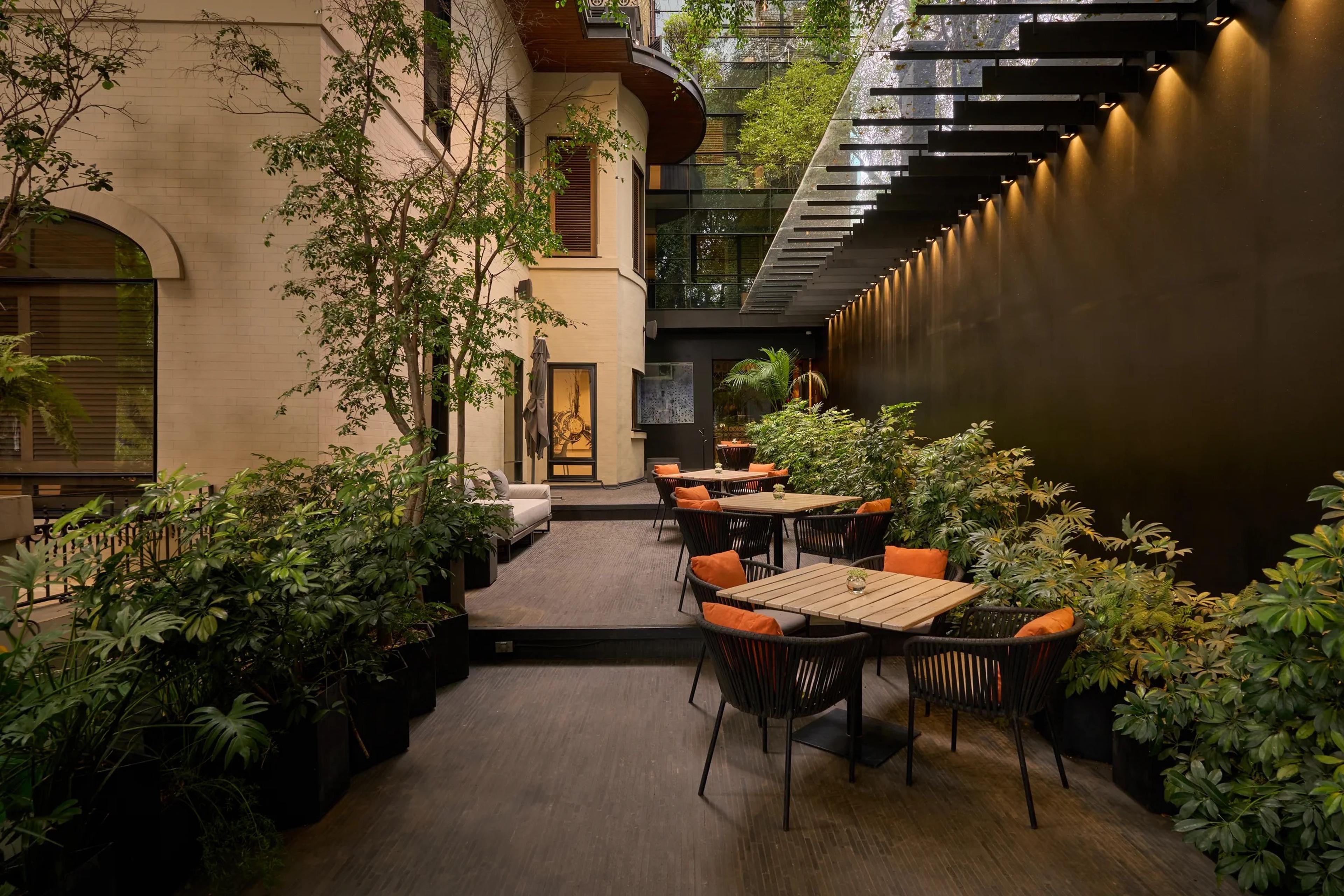 A dusk view of the hotel's cream facade and arched windows beside an outdoor terrace with orange cushioned chairs, trees and a lit stairway.