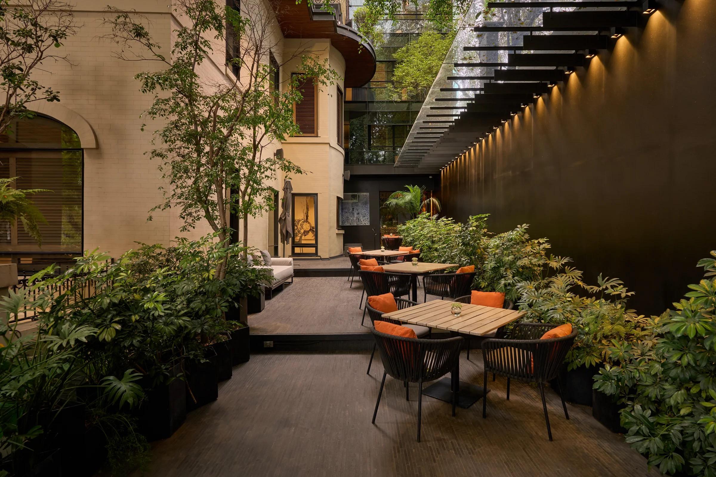 A dusk view of the hotel's cream facade and arched windows beside an outdoor terrace with orange cushioned chairs, trees and a lit stairway.