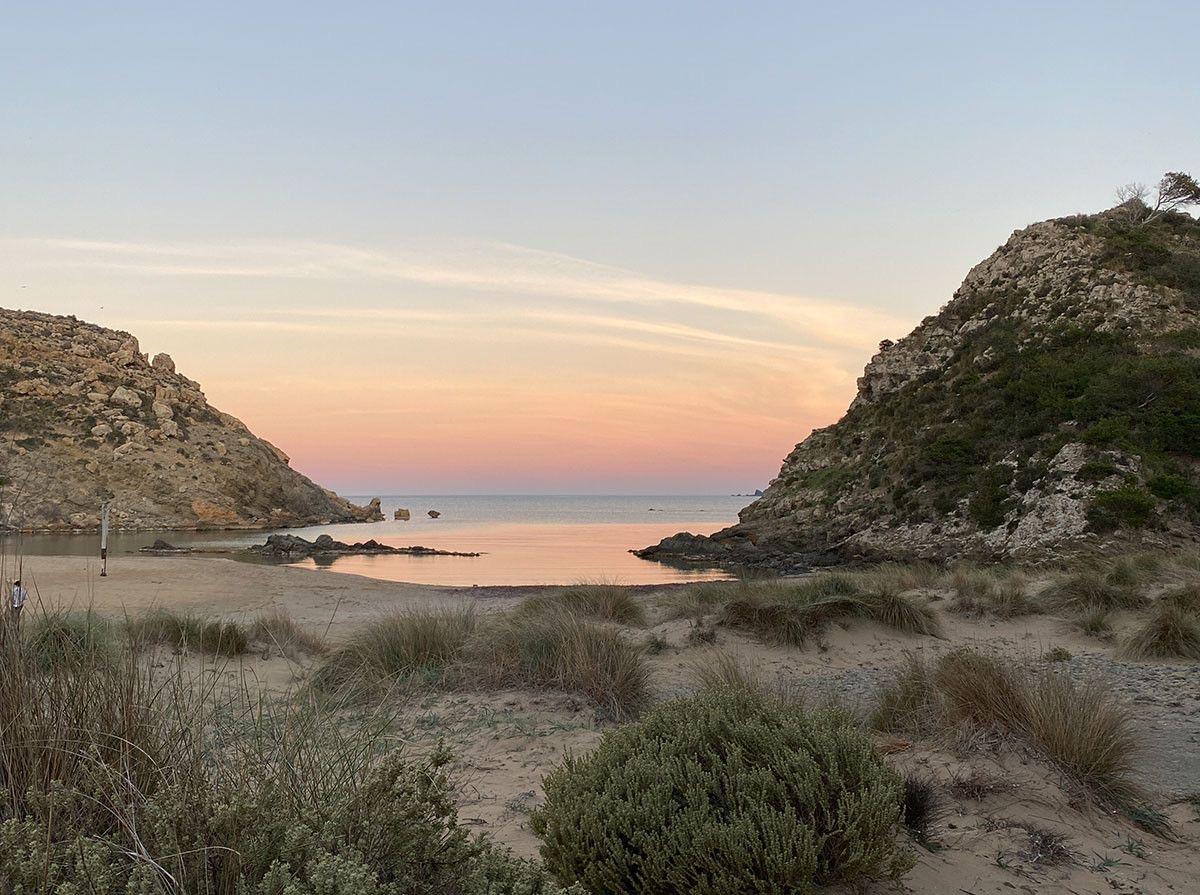 A rocky cove at dusk, pink sky over still water between two headlands, with one small figure far off on the sand.