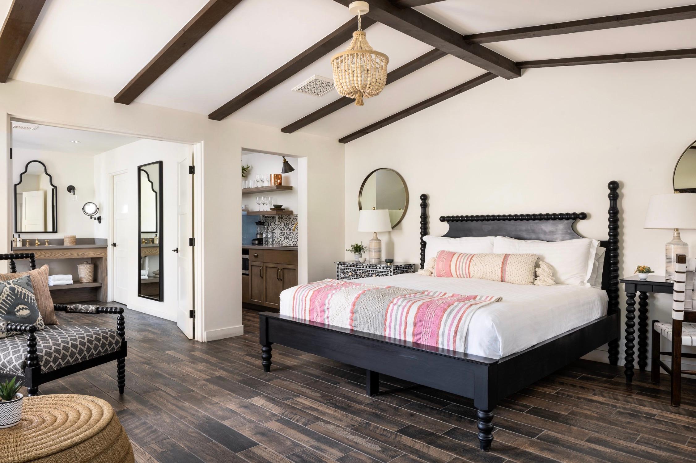 A bedroom with a black spindle bed dressed in white linen and a striped throw, a beaded chandelier, exposed wood beams and dark wood floors.