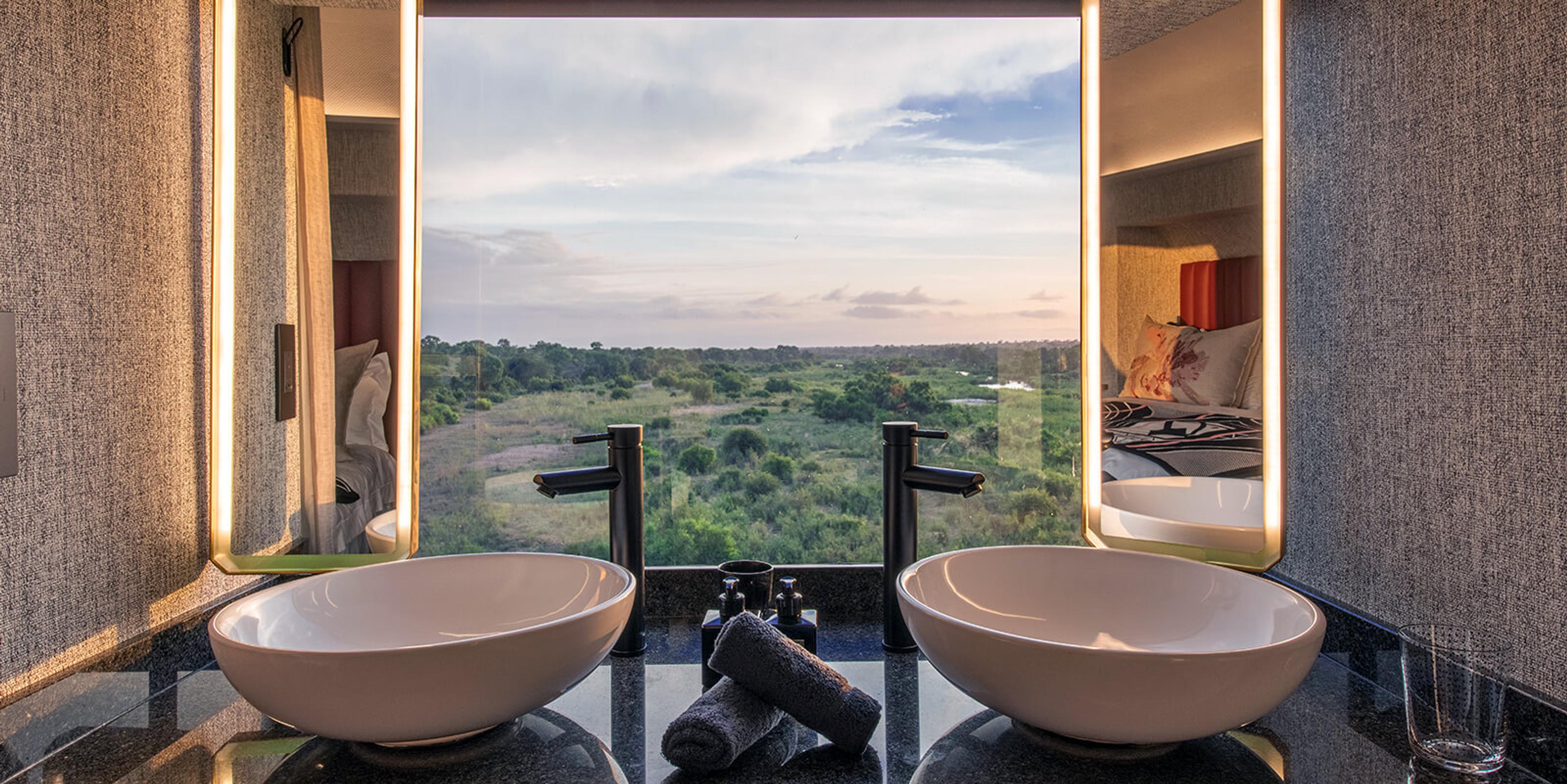 Two round basin sinks in a bathroom, framed by a wide window looking straight out over open bushveld.