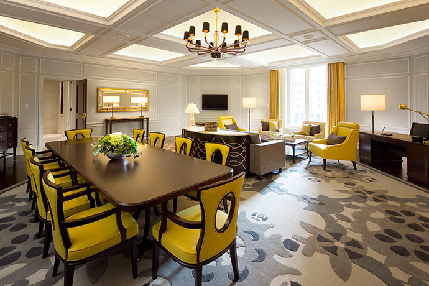 A spacious hotel suite with a long dining table and ten yellow chairs, a seating area with armchairs and a television, gold curtains at tall windows, and a patterned grey carpet.
