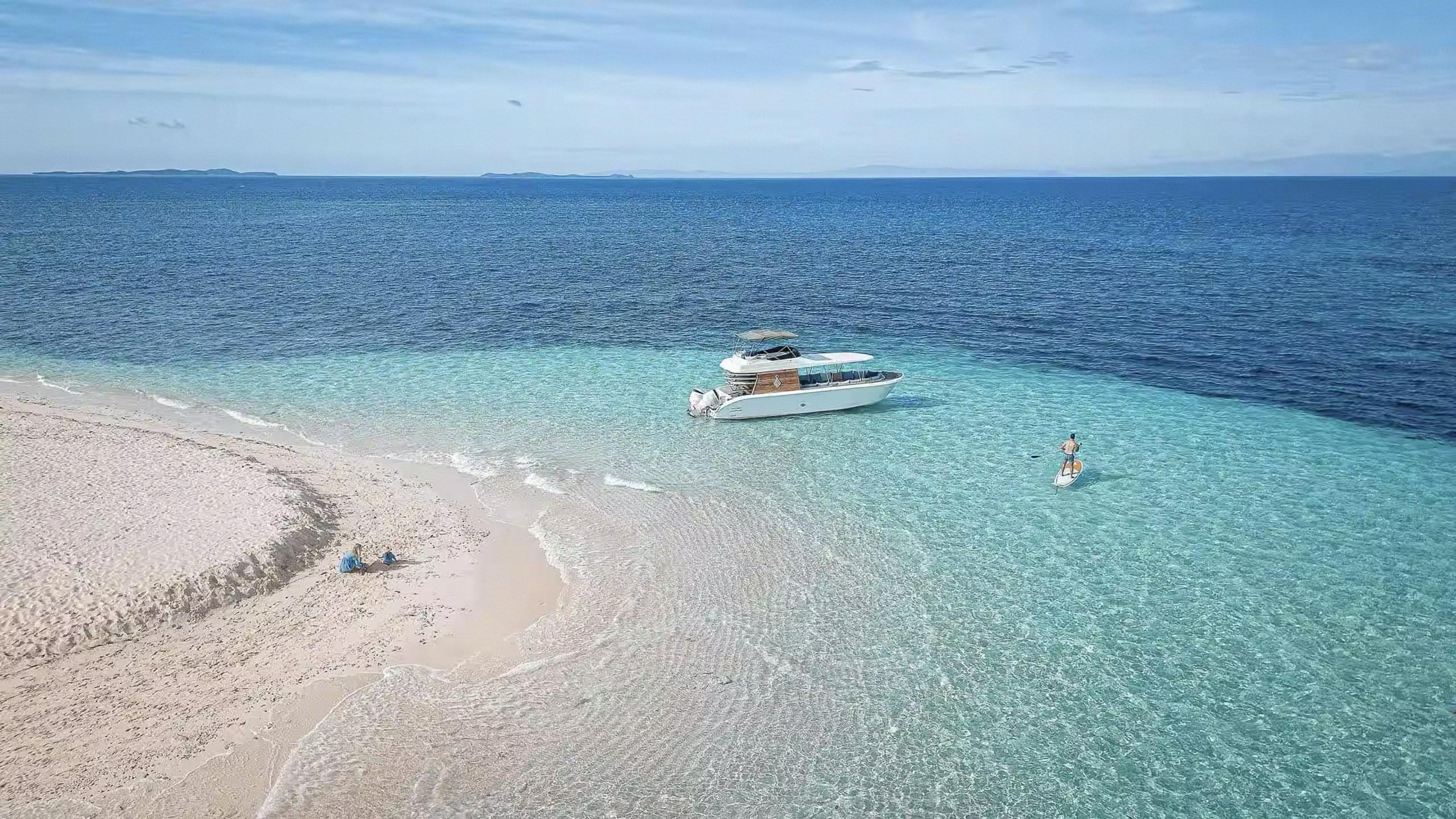 A speedboat and a person on a paddleboard anchored off a white sandbar in turquoise water, with two people sitting on the sand nearby.