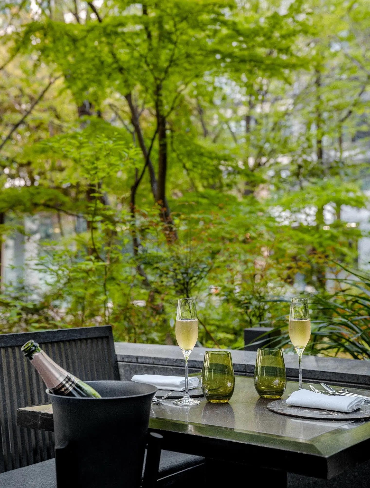 A table set on an outdoor terrace with two glasses of champagne and a bottle in an ice bucket, surrounded by trees.