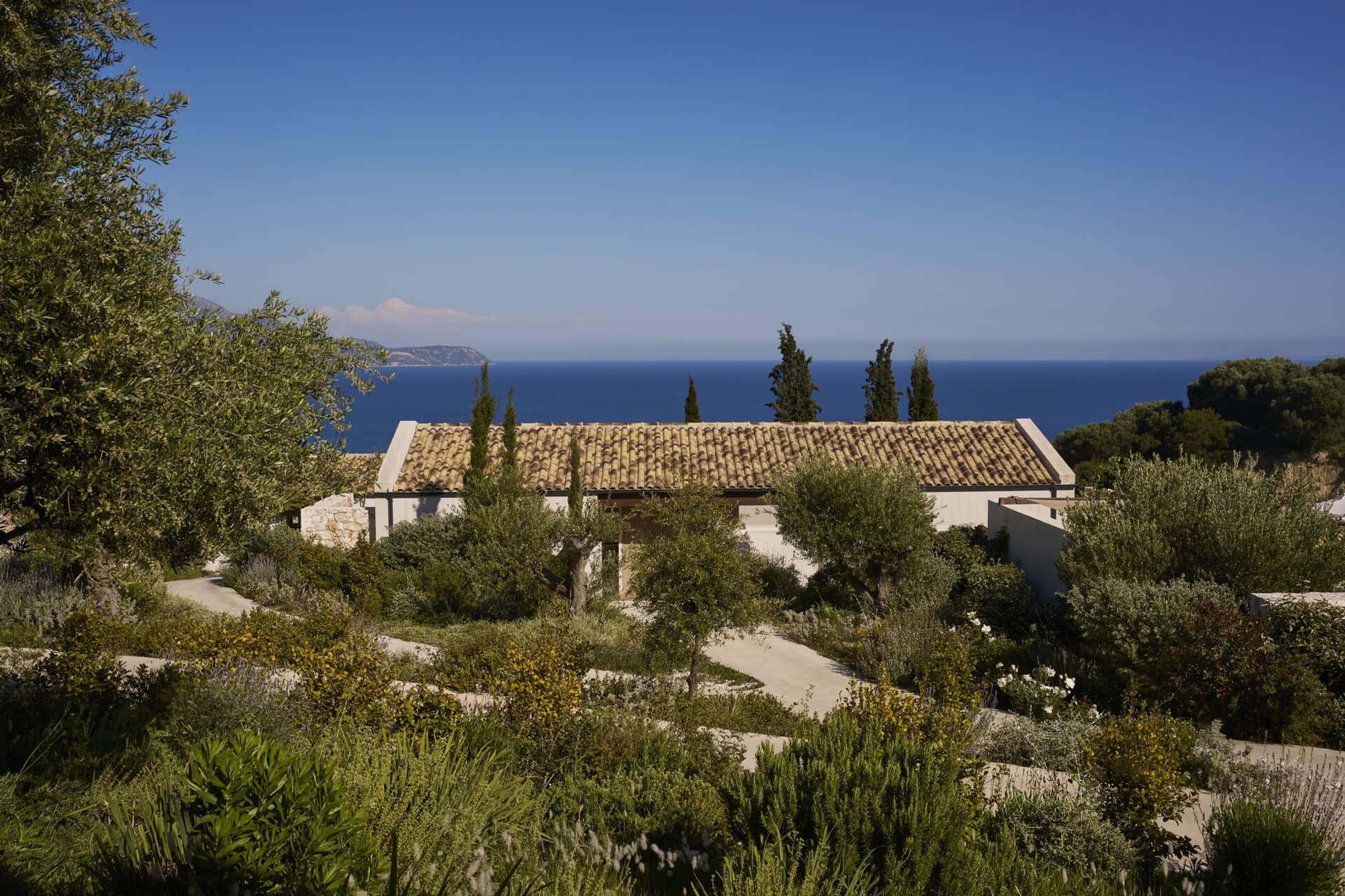 a low white and stone building with a terracotta roof, set among trees and shrubs with the sea filling the horizon behind it